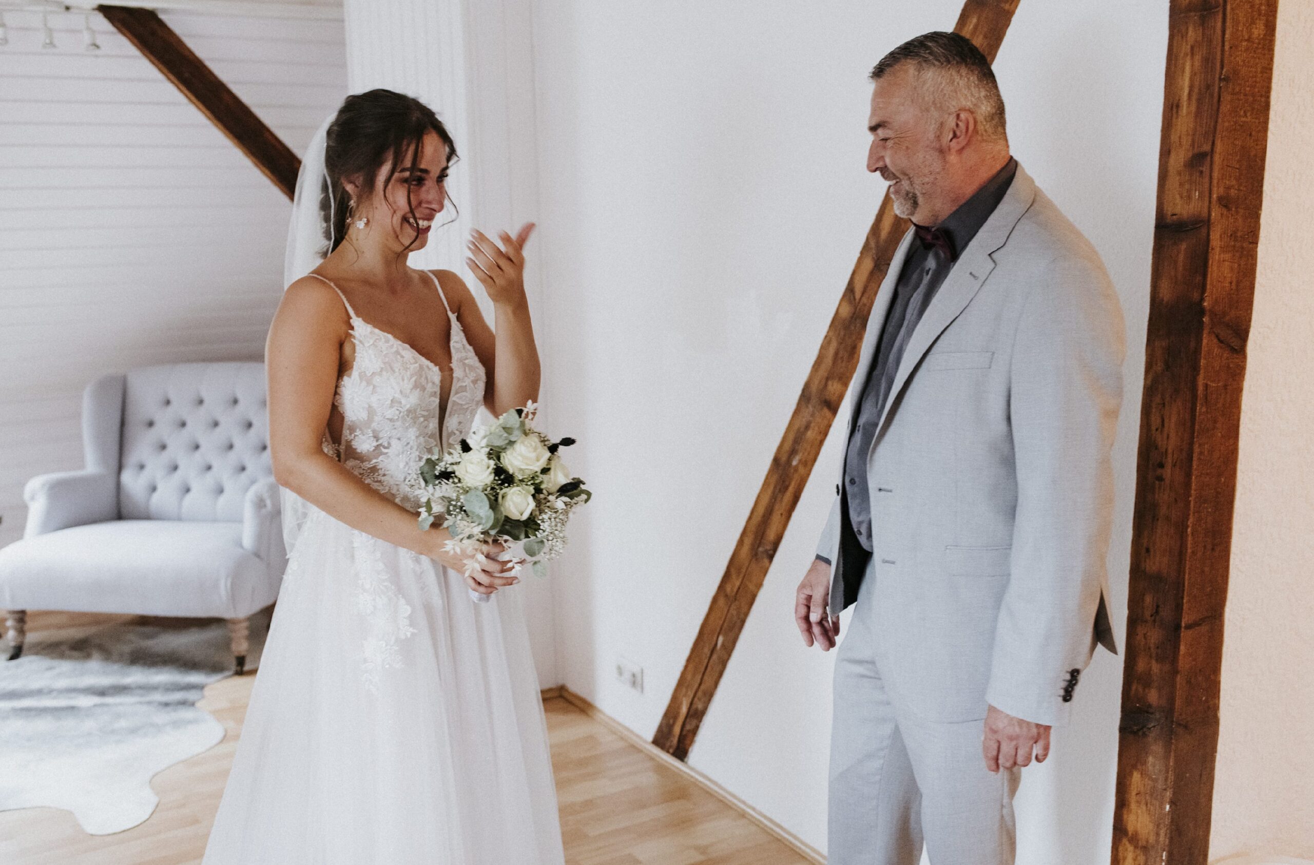 Braut im weißen Kleid mit Blumenstrauß neben älterem Mann im Anzug, Holzbalken im Raum.