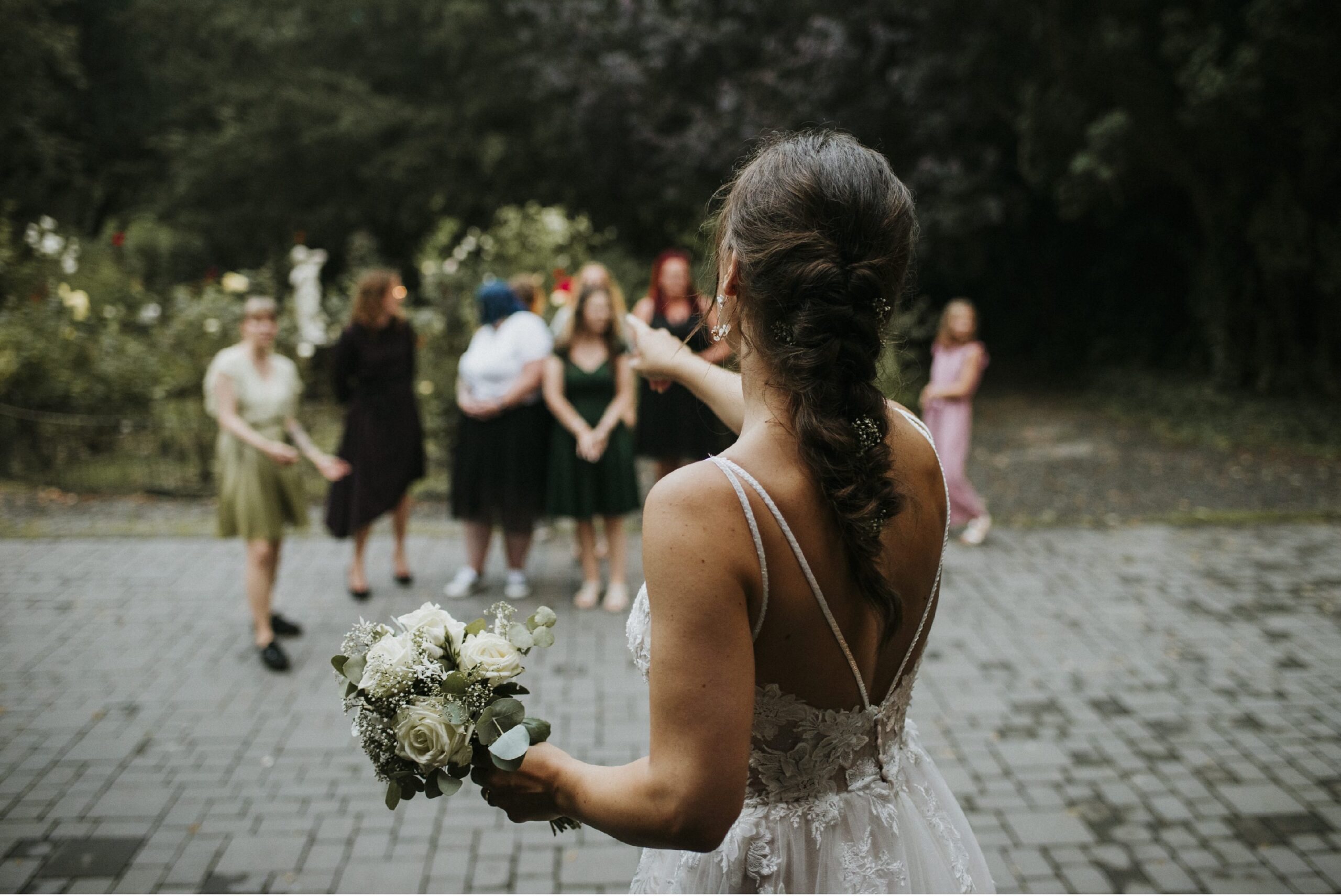 Braut im Hochzeitskleid mit Blumenstrauß, wendet sich einer Gruppe im Freien zu.