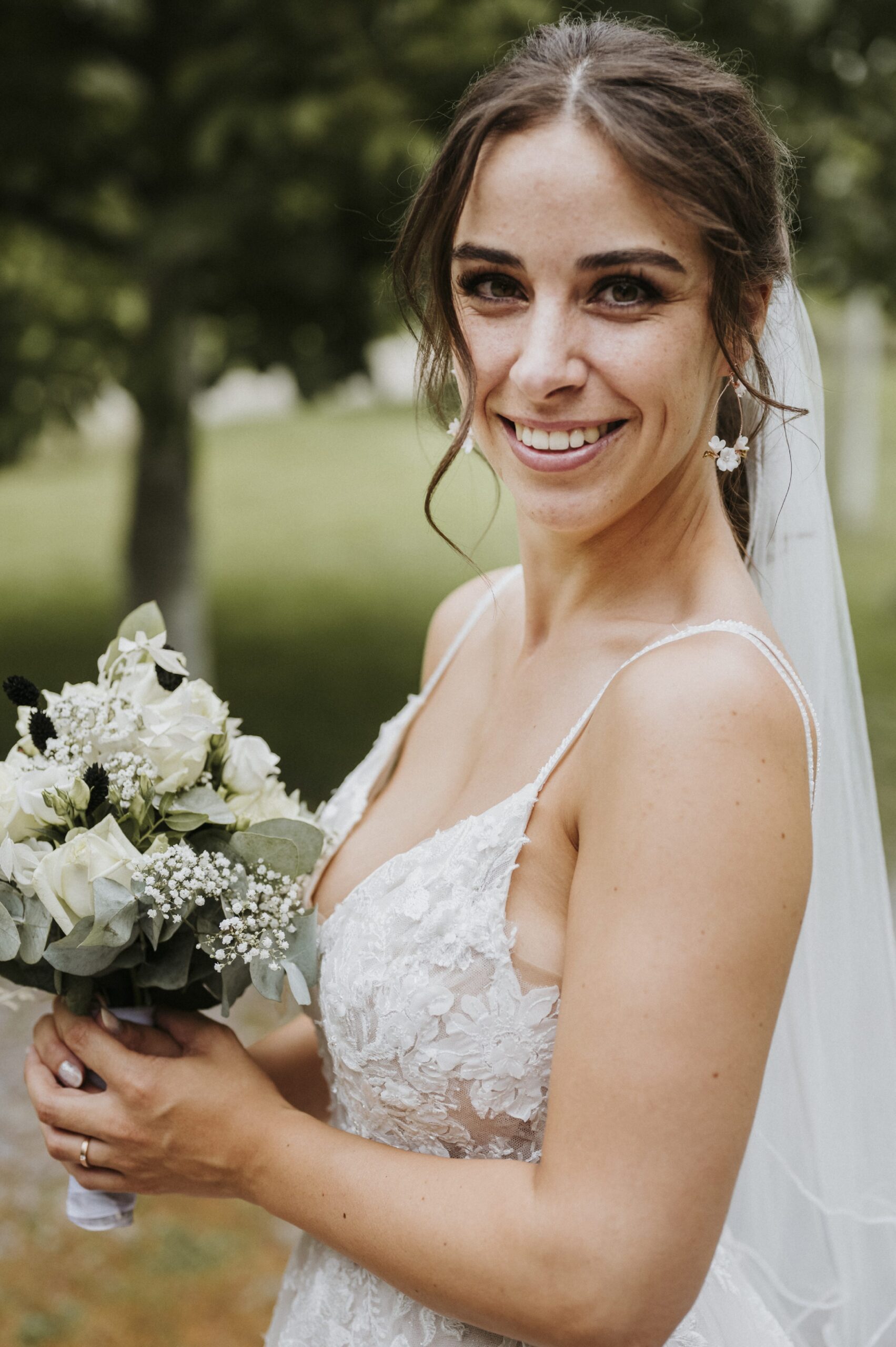 Braut im weißen Spitzenkleid mit Schleier und Blumenstrauß, lächelnd im Freien vor Bäumen.