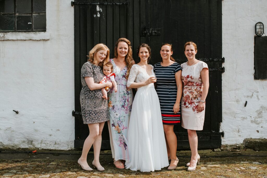 Fünf Frauen vor einer weißen Wand mit schwarzer Tür, eine trägt ein Brautkleid, eine ein Baby.