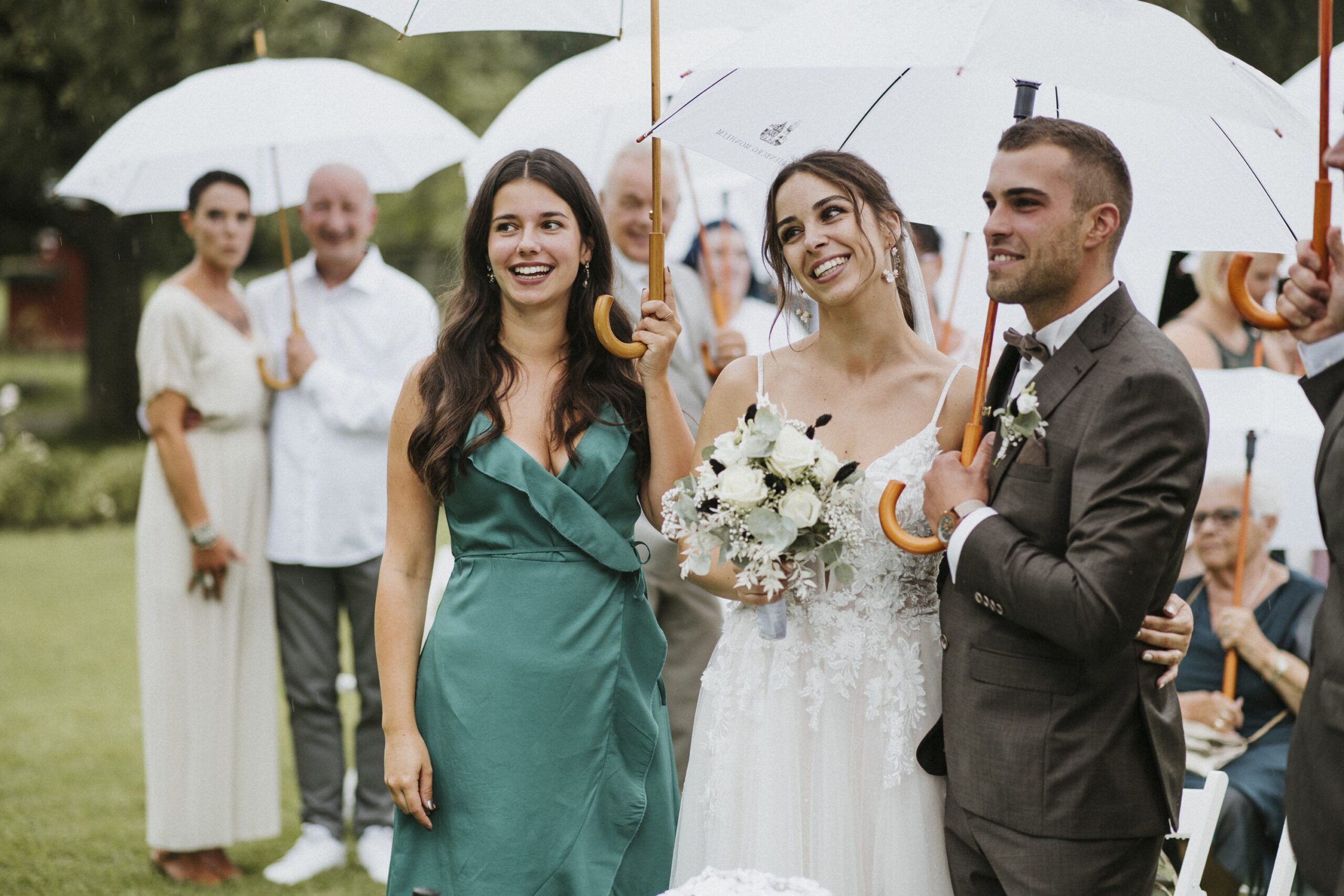 Brautpaar und Frau unter Regenschirmen bei Hochzeit im Freien mit Blumenstrauß.
