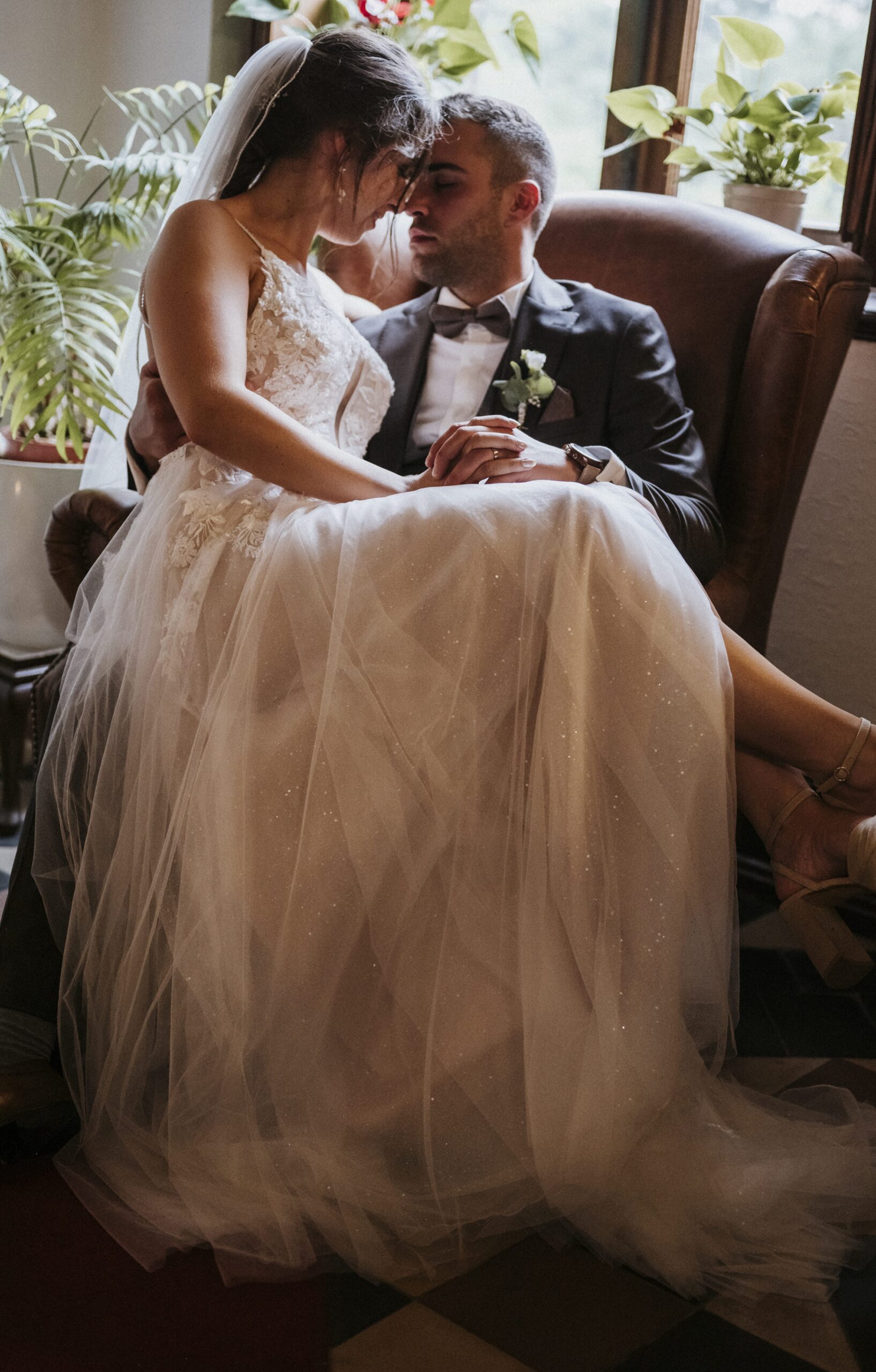 Brautpaar in Hochzeitskleidung auf Ledersessel, Pflanzen im Hintergrund.