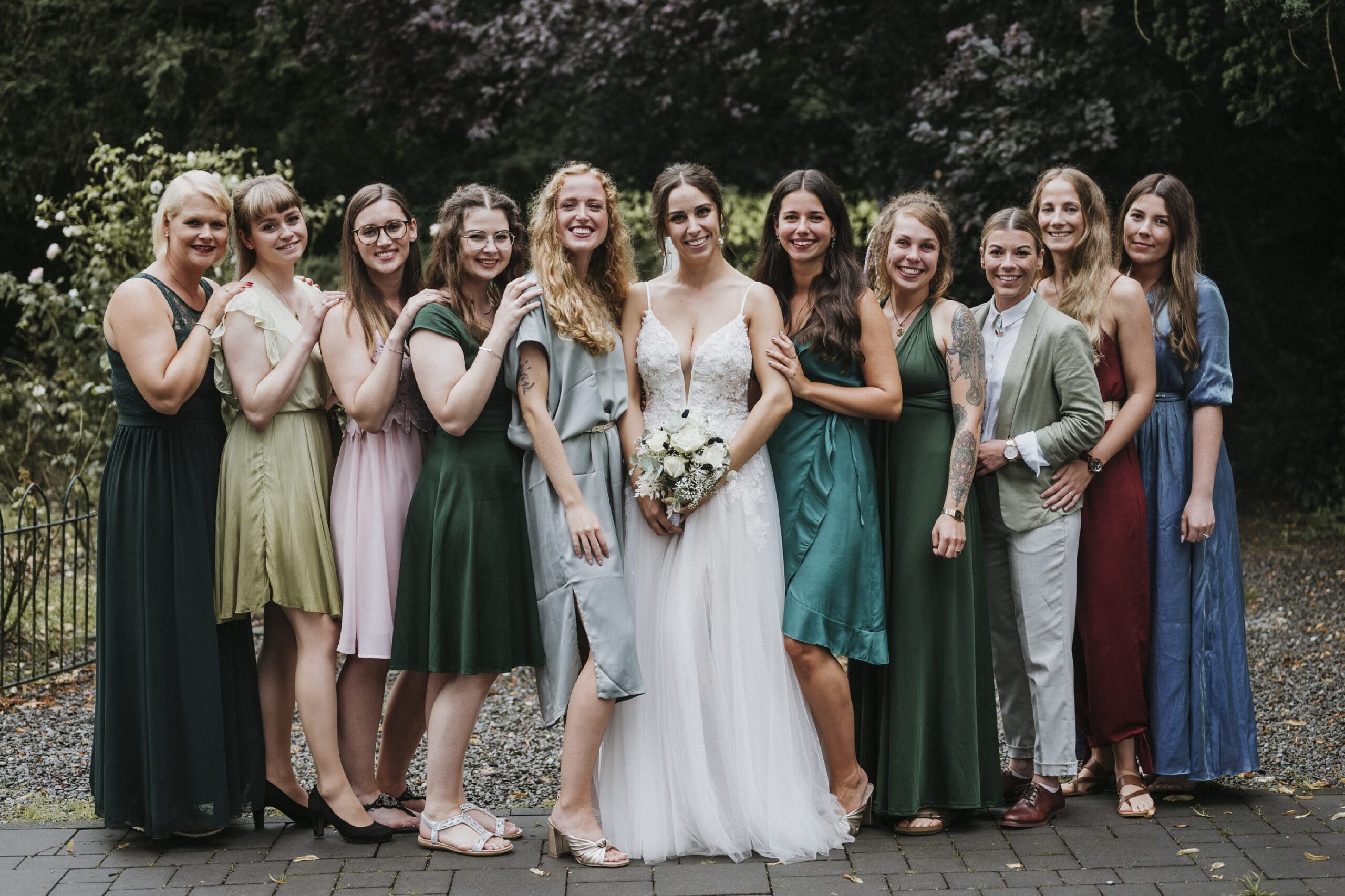 Eine Braut in Weiß mit elf Frauen in bunten Kleidern posiert draußen für ein Foto.