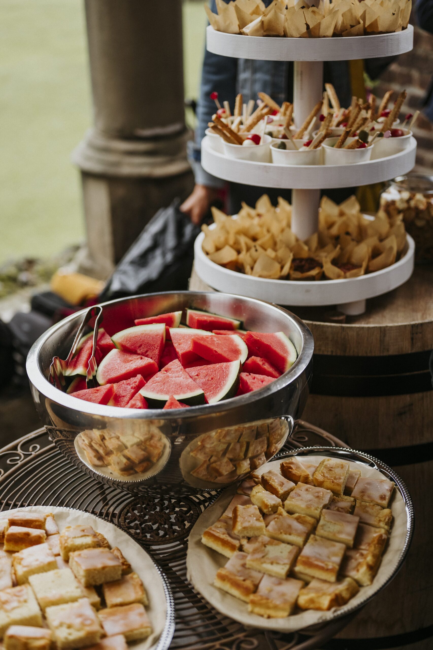 Buffet mit Wassermelone, Gebäck und dreistufigem Servierständer auf einem Tisch im Freien.