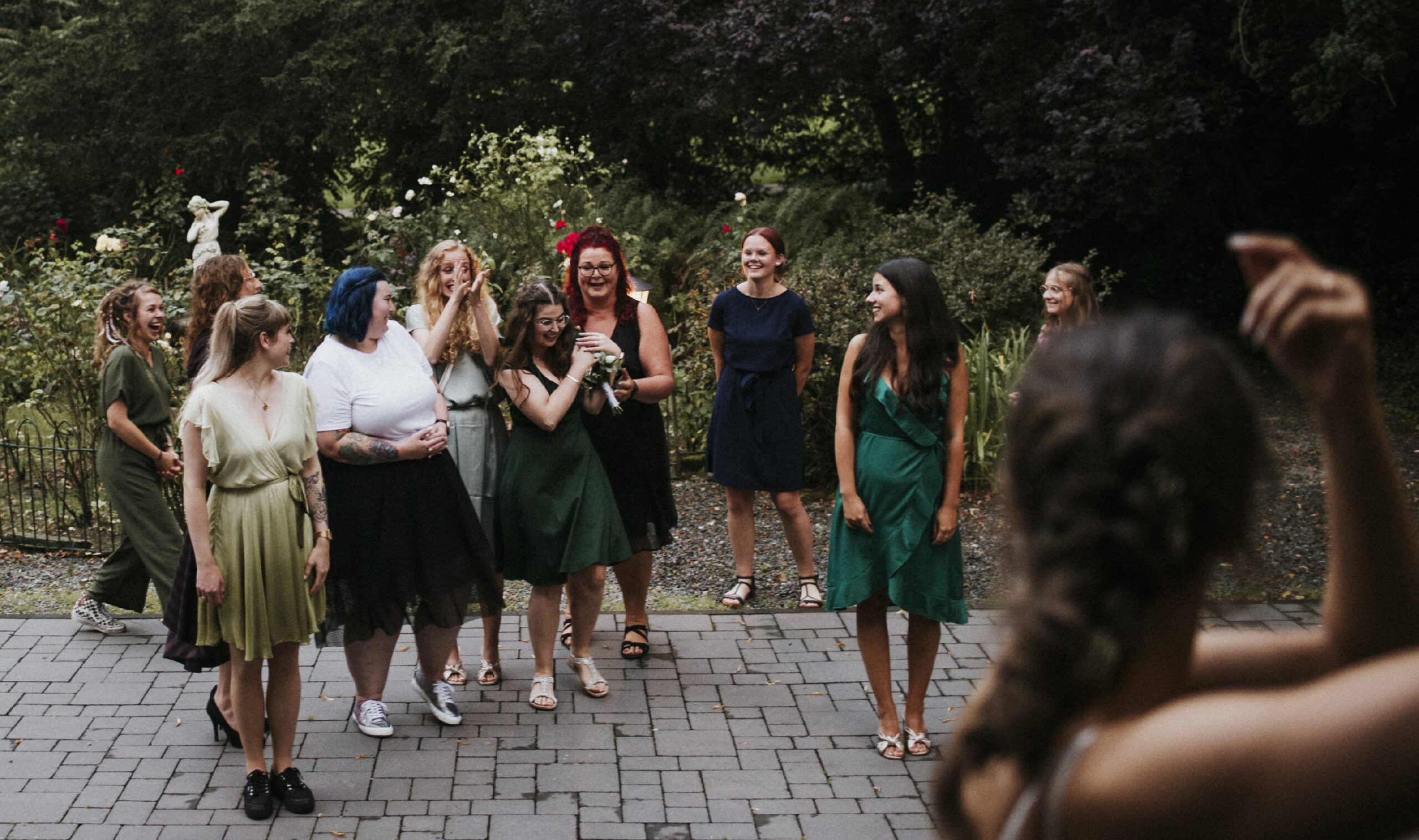 Frauen in eleganten Kleidern auf gepflastertem Weg im Garten, lachend vor grünen Büschen.