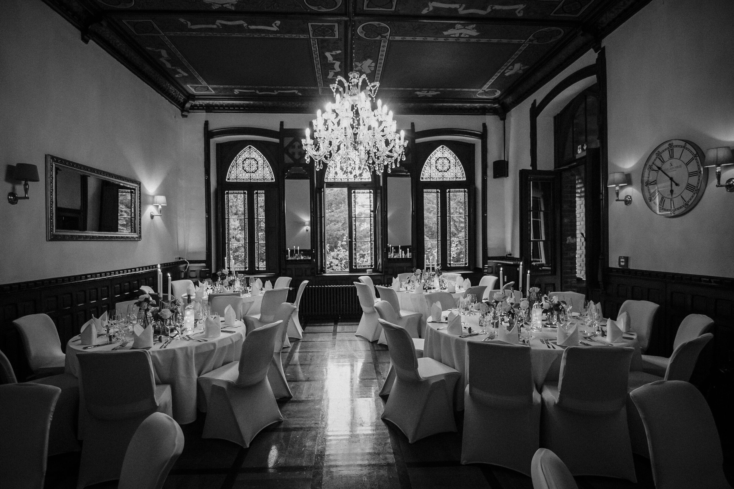 Eleganter Speisesaal mit runden Tischen, weißen Stuhlhussen, Kronleuchter, großen Fenstern.
