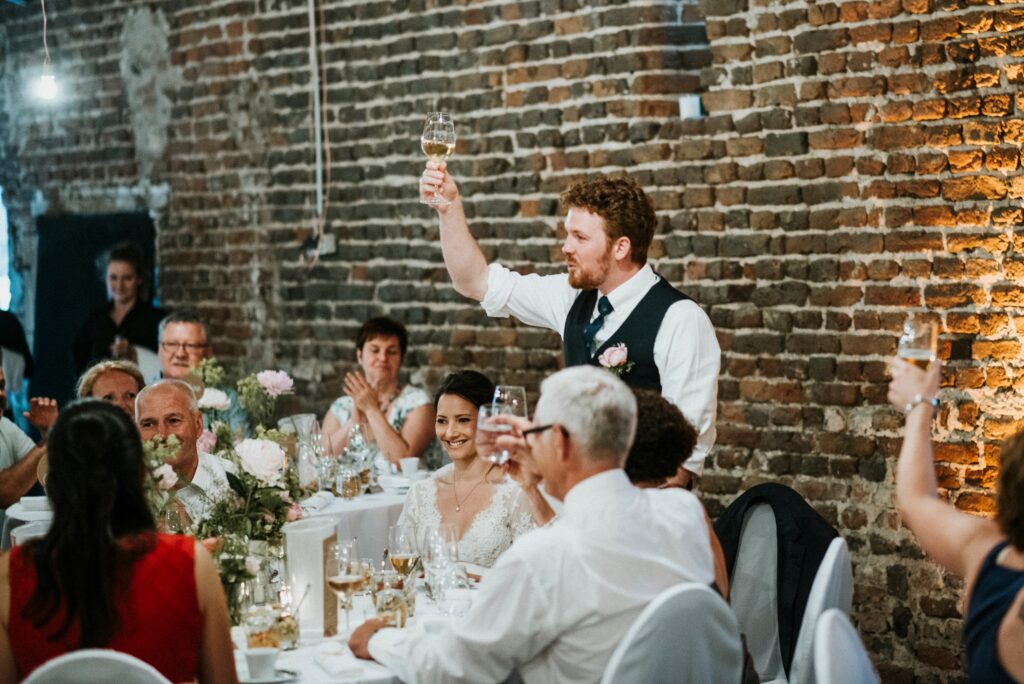 Person toasts at a festive event with guests applauding, brick wall in the background.
