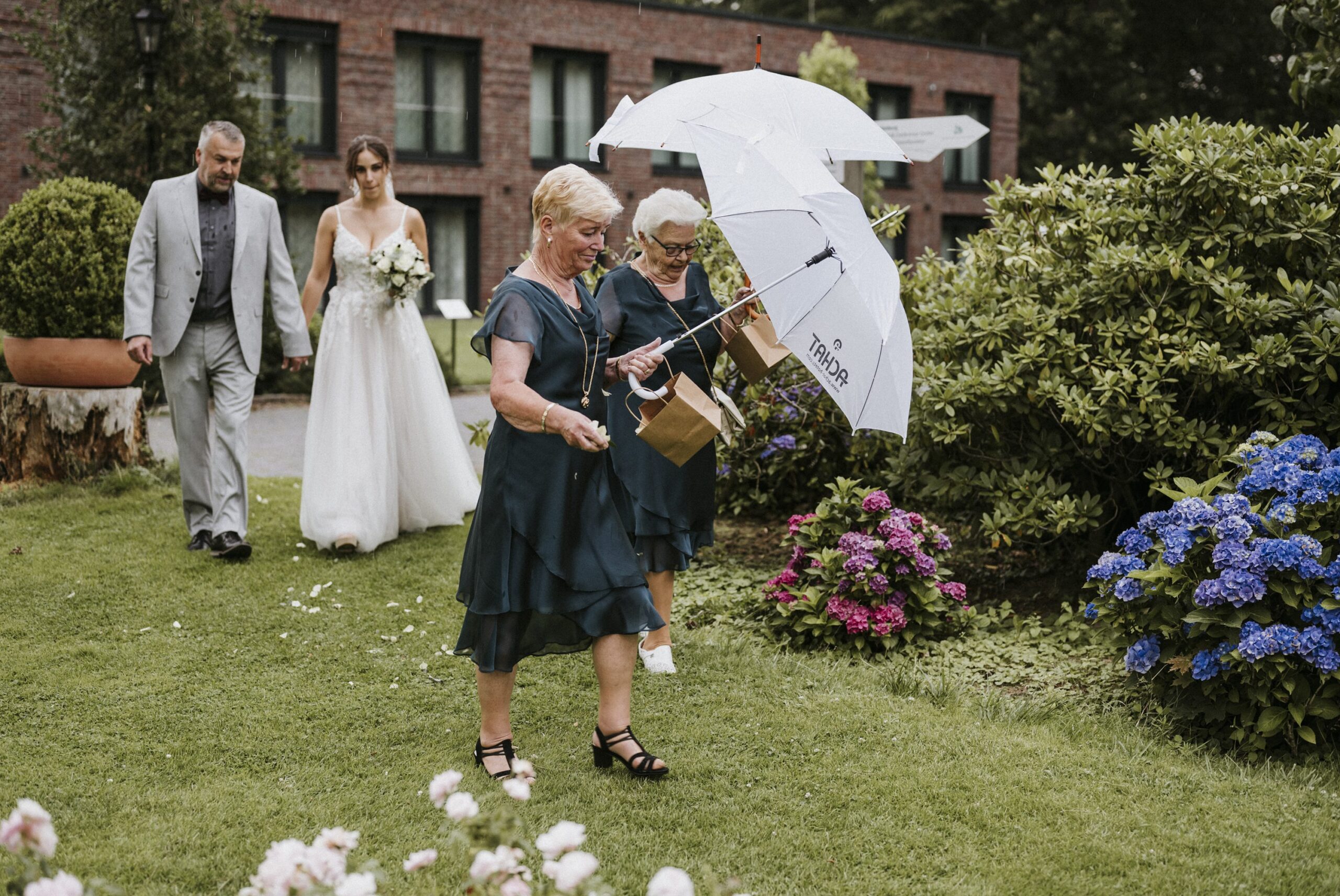 Eine Frau im weißen Kleid und ein Mann folgen zwei Frauen mit Regenschirmen auf grünem Rasen.