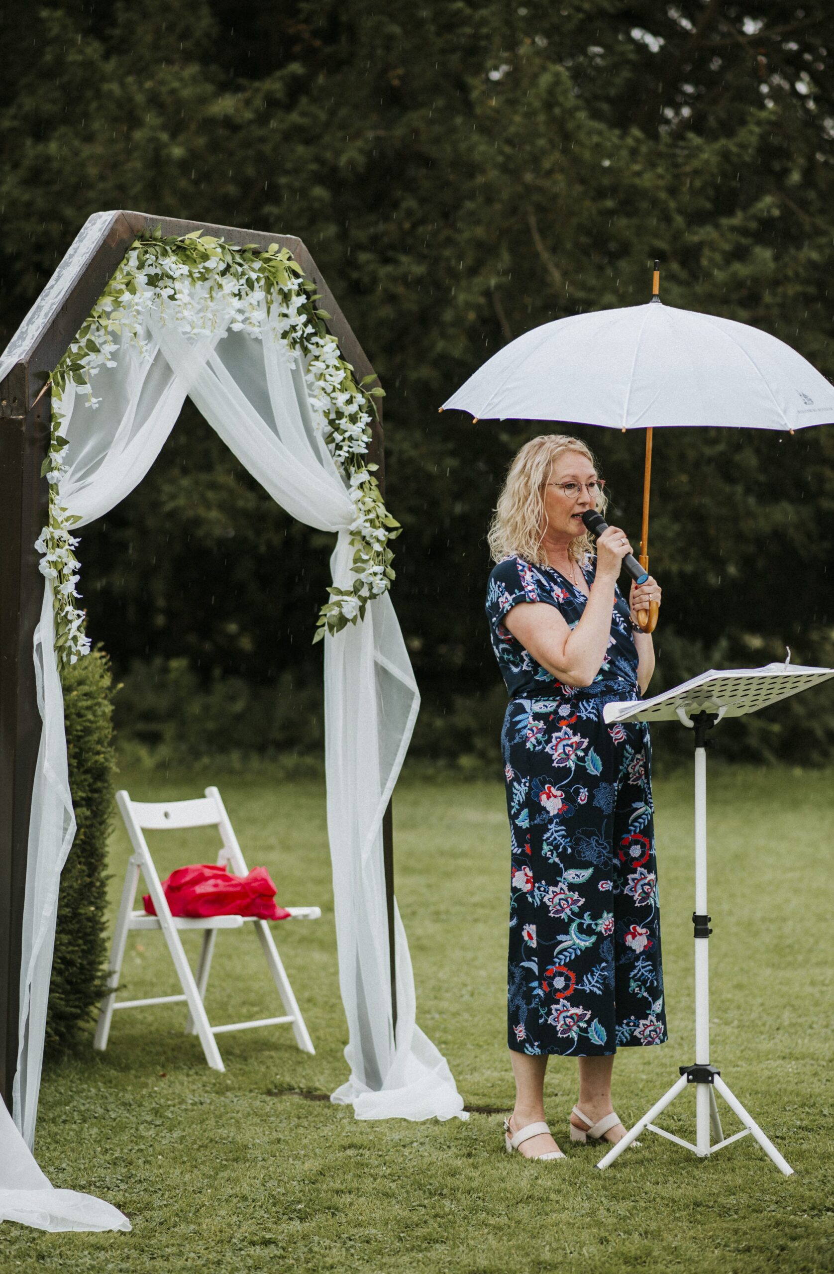 Frau hält Rede unter Regenschirm vor Traubogen mit Tüchern und Blumen auf Wiese.