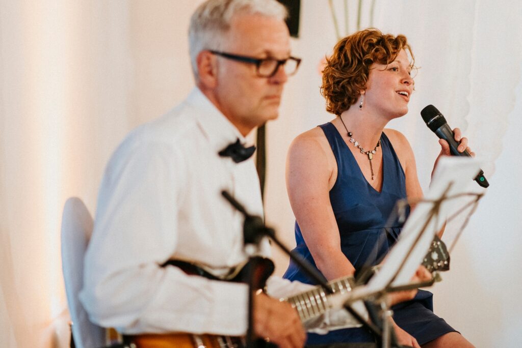 Frau in blauem Kleid singt, Mann spielt Gitarre, beide elegant gekleidet im Innenraum.