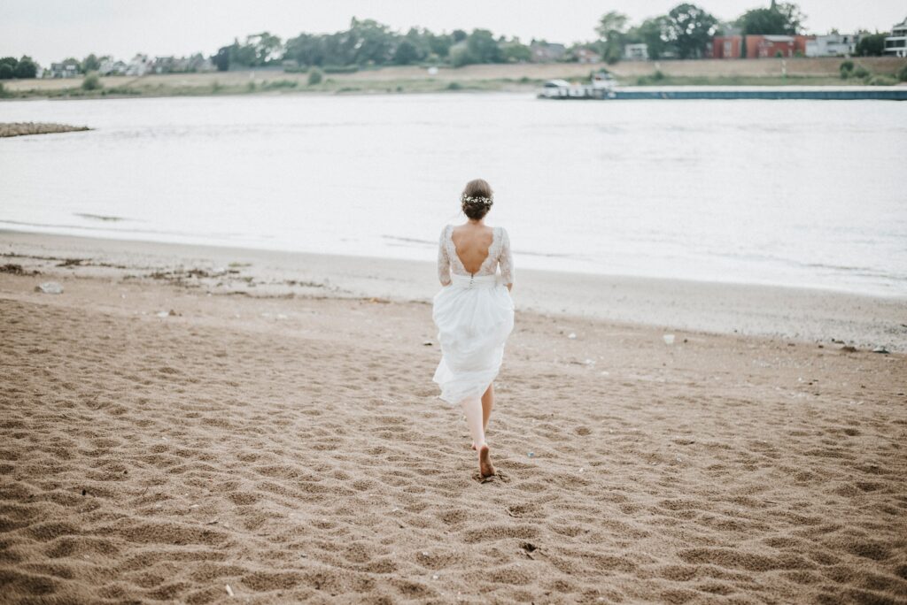 Frau im weißen Kleid läuft am Strand zum Wasser; Bäume und Gebäude im Hintergrund.