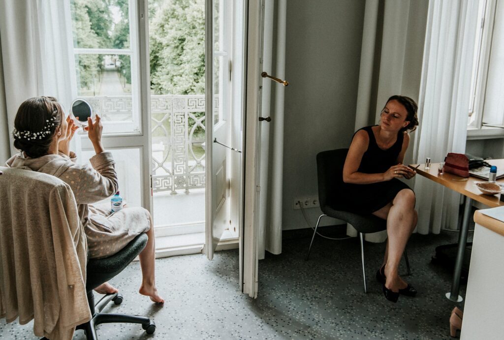 Zwei Frauen in hellem Raum, eine am Tisch schminkt sich, die andere hält Spiegel am Fenster.