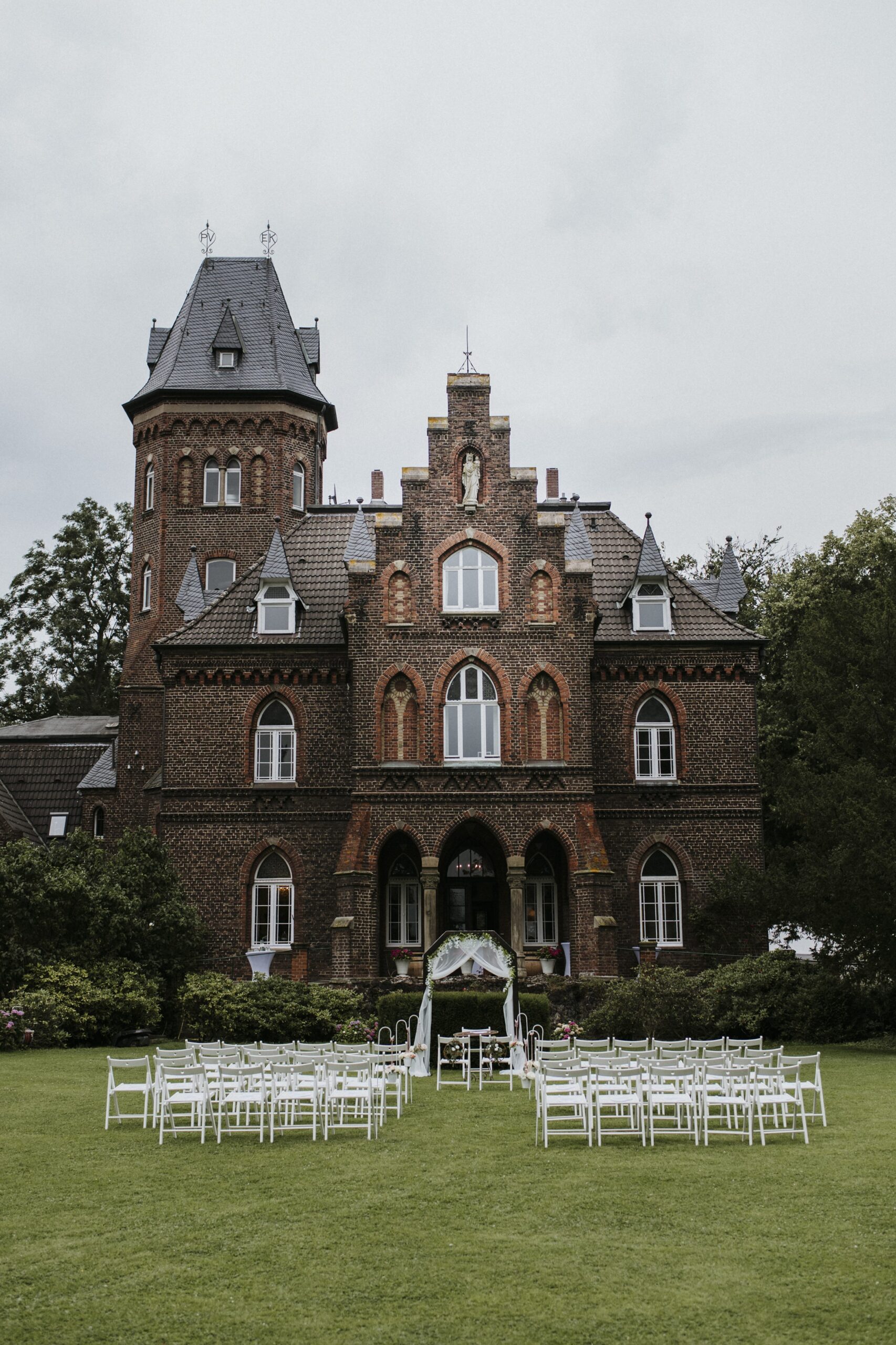 Historisches Backsteingebäude mit Turm, Rasenfläche mit weißen Stühlen und dekoriertem Torbogen.