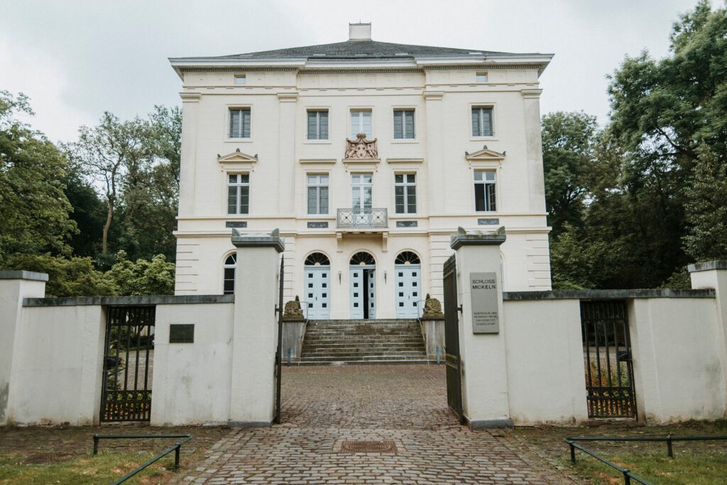 Historisches Gebäude mit symmetrischer Fassade, Bäumen, Kopfsteinpflaster und offenem Tor.