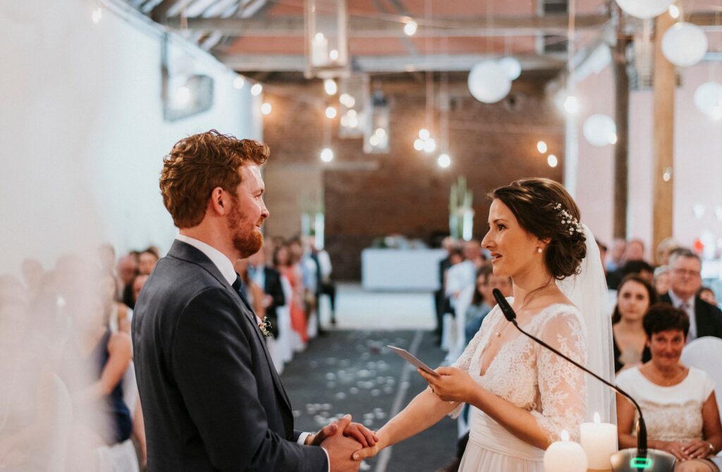Hochzeitspaar hält Hände; die Braut spricht ins Mikrofon vor sitzendem Publikum im festlichen Raum.