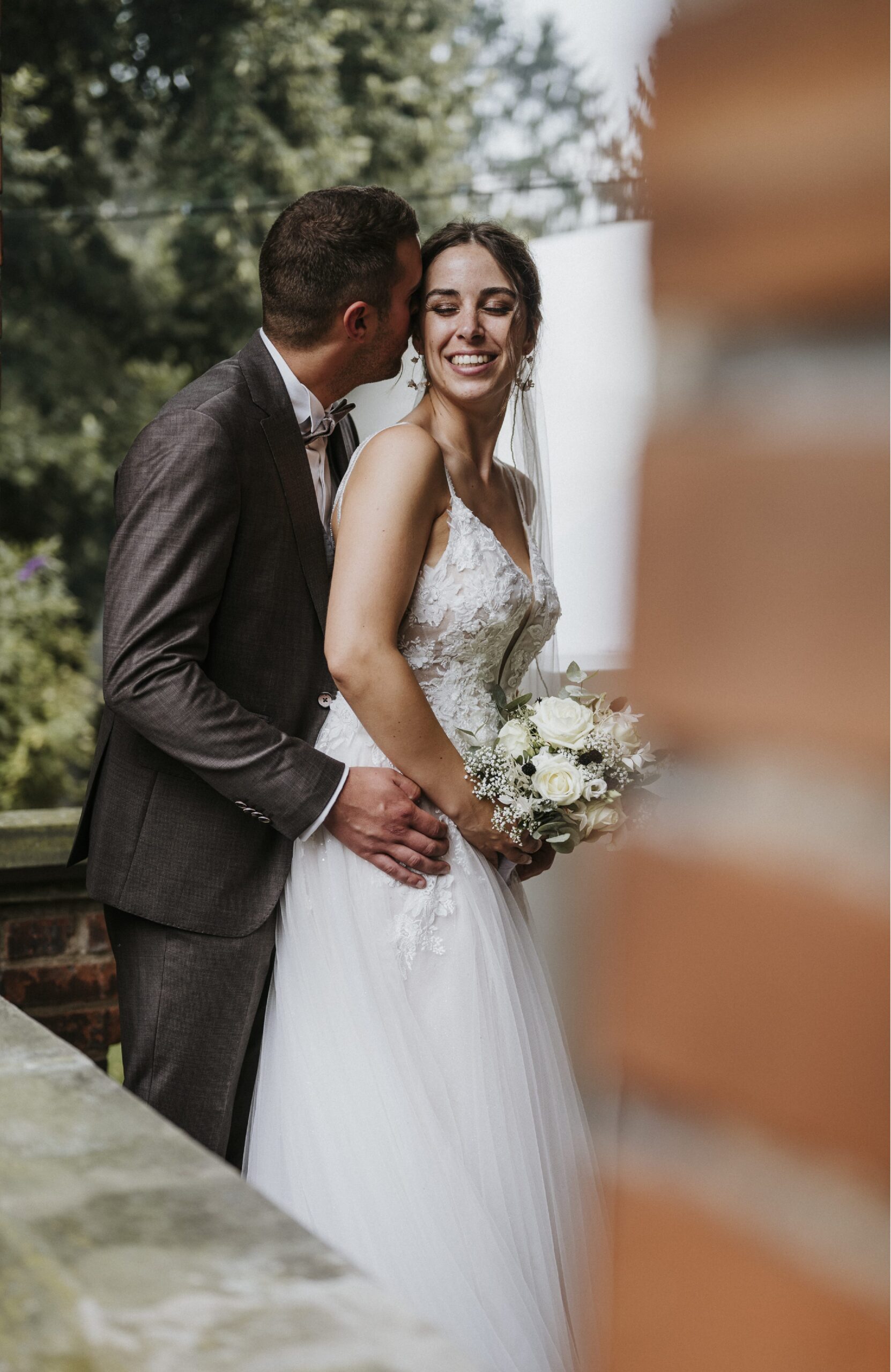 Hochzeitspaar auf Balkon, Braut mit Blumenstrauß, Bräutigam im Anzug, unscharfe Bäume im Hintergrund.
