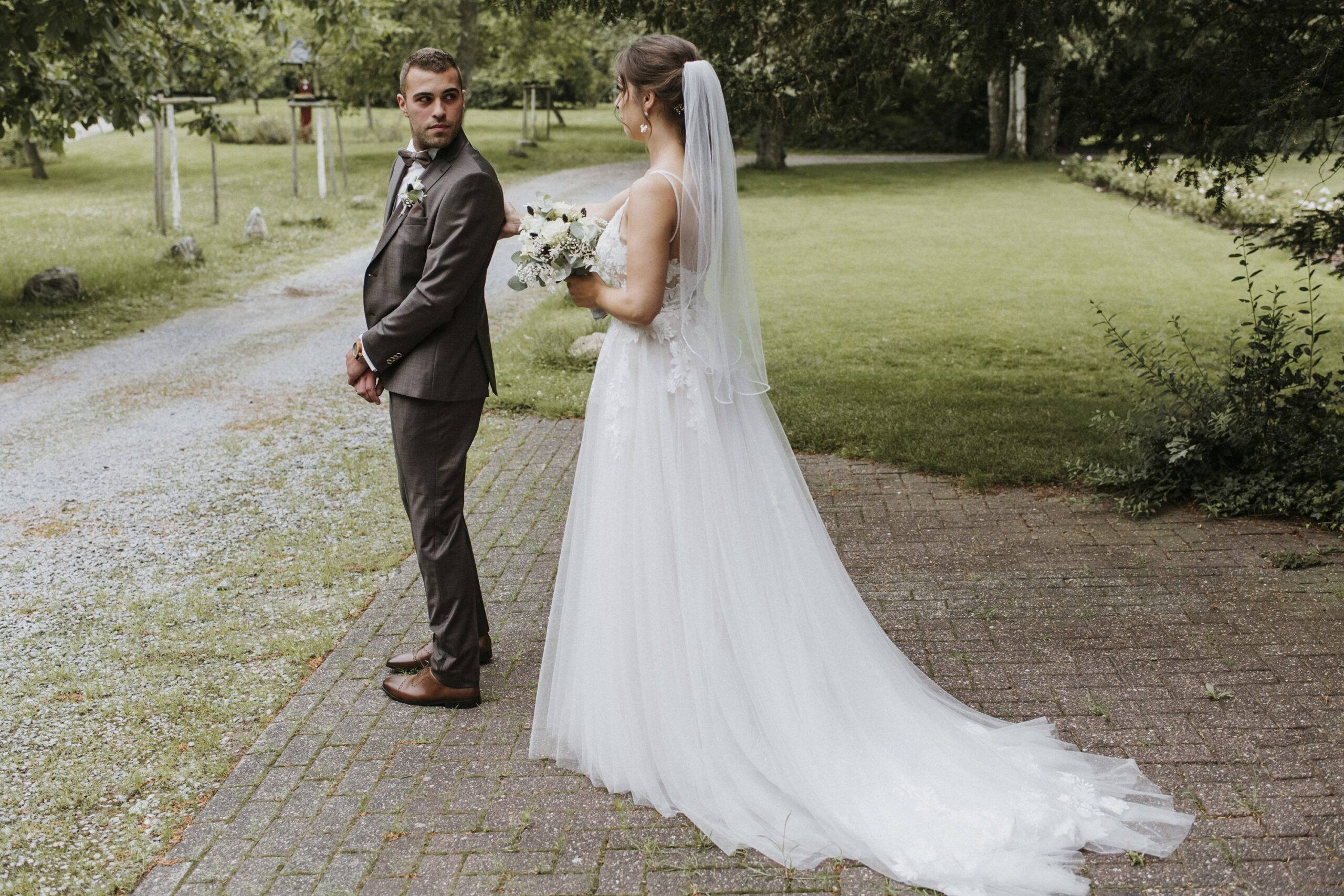 Hochzeitspaar im Freien, Braut in weißem Kleid mit Schleier, Bräutigam im Anzug, grüne Kulisse.