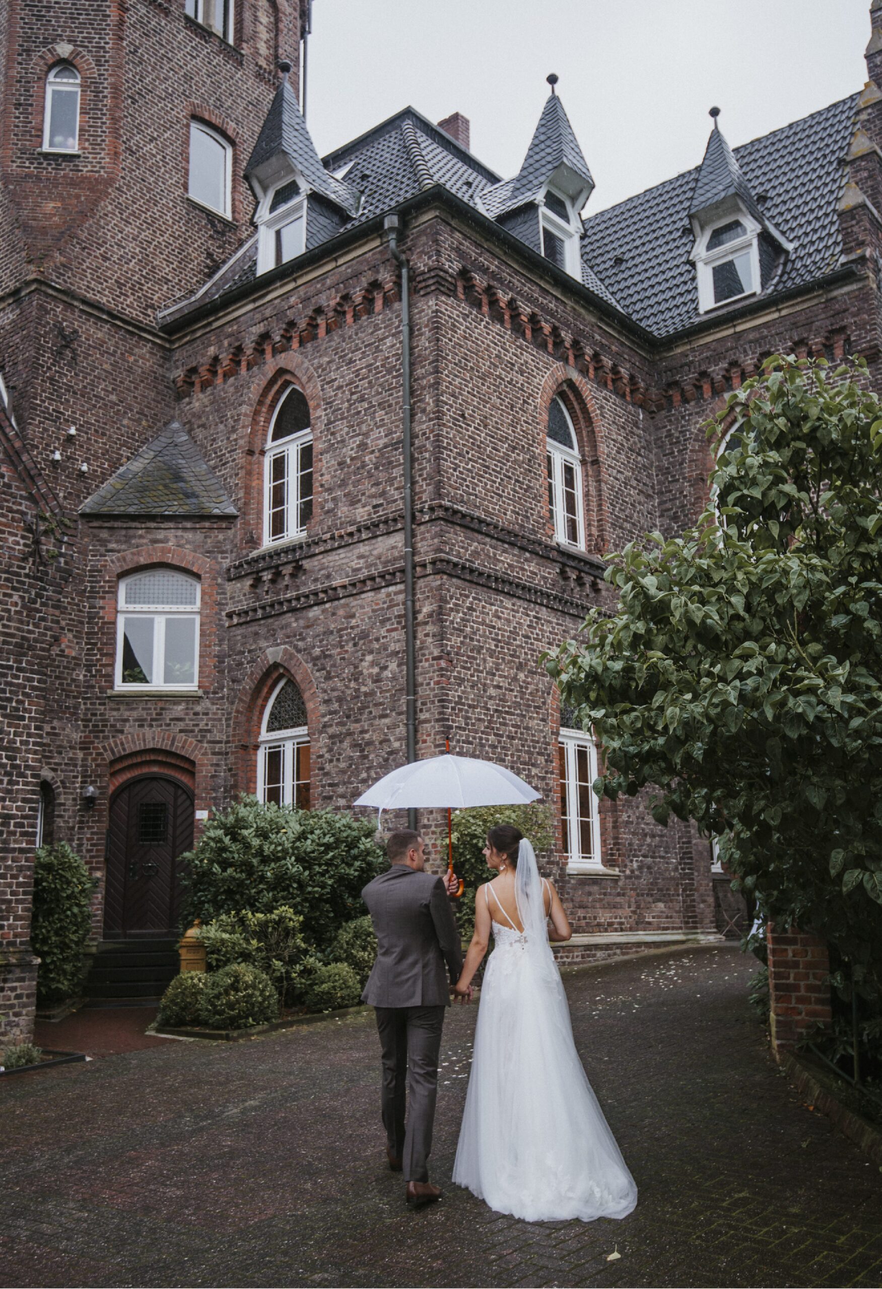 Hochzeitspaar mit Regenschirm vor historischem Backsteingebäude und grünen Sträuchern.