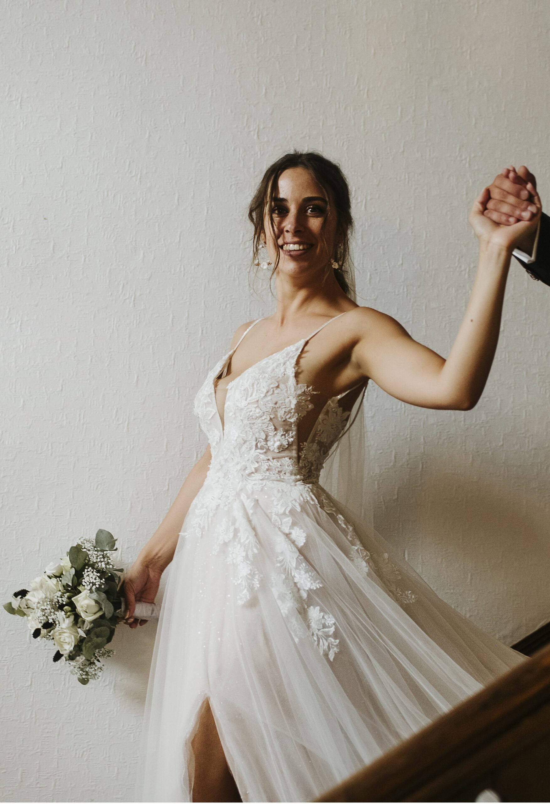 Braut lächelnd im ärmellosen Kleid mit Blumenstrauß, hält Hand, auf Treppe.