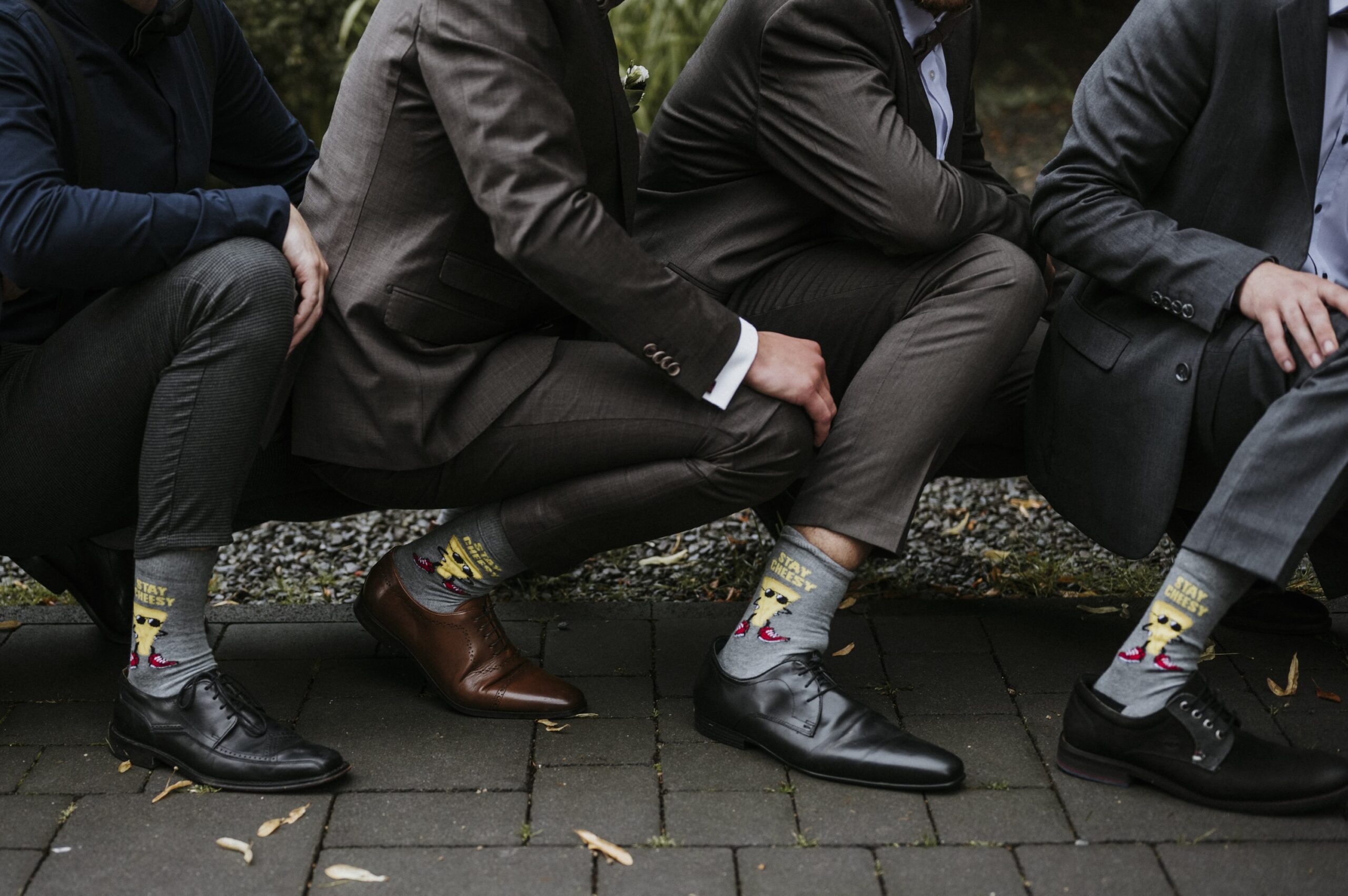 Männer in Anzügen hocken, tragen bunte Comicsocken und Lederschuhe auf Gehweg.