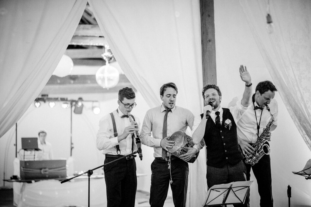Vier Männer in Anzügen musizieren mit Flöte, Trommel, Mikrofon, Saxophon; DJ im Hintergrund.