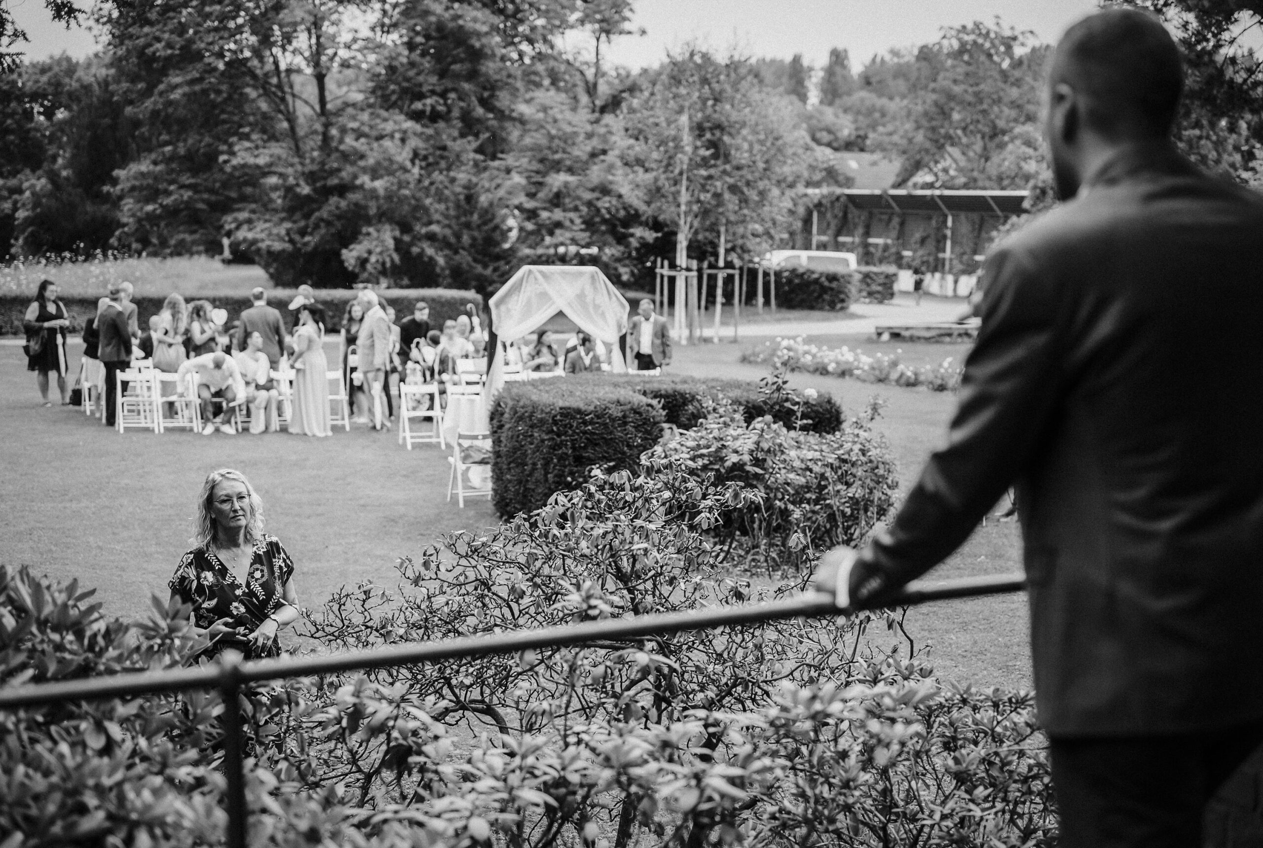 Mann auf Plattform beobachtet Frau, hinten Gruppe bei Veranstaltung in parkähnlicher Umgebung