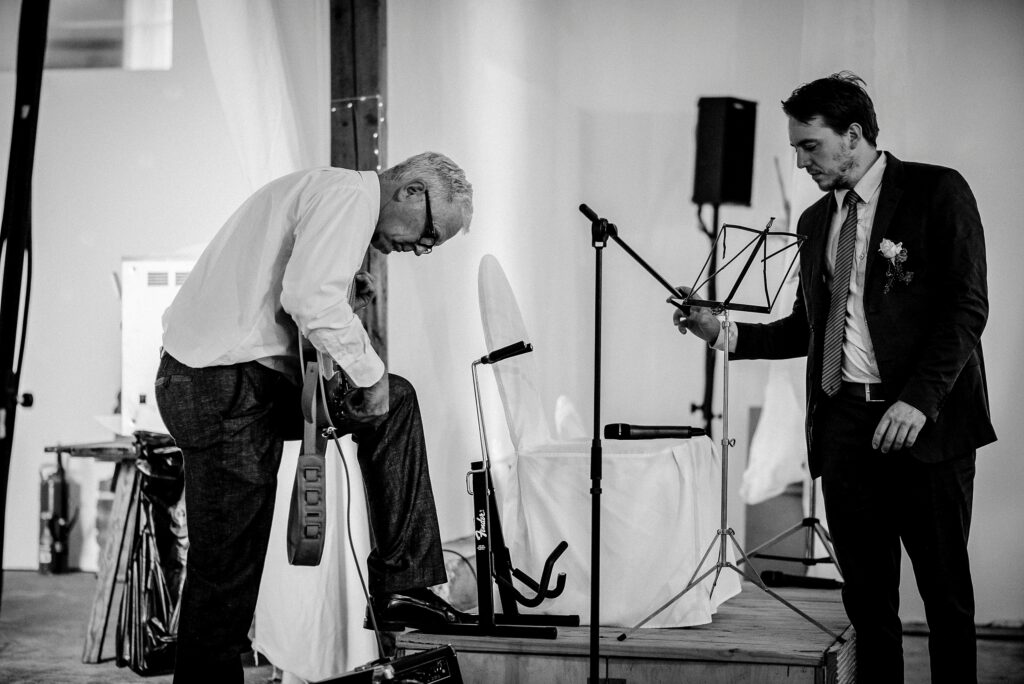 Mann mit Gitarre tritt auf Pedal, anderer passt Notenständer an, Mikrofon im Hintergrund.