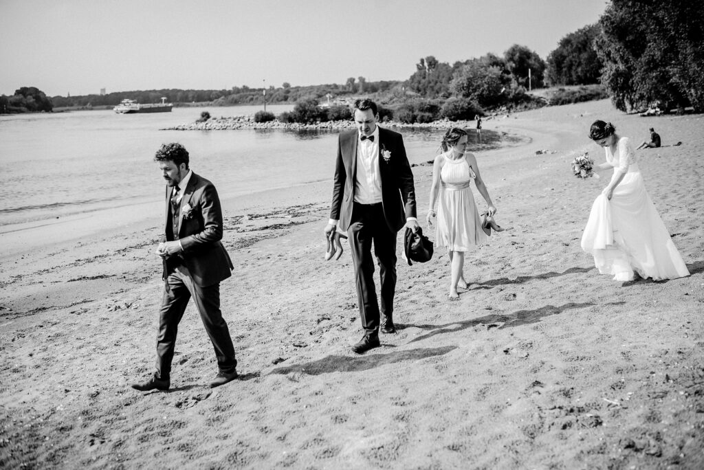 Gruppe von Menschen in formeller Kleidung barfuß am Strand mit Fluss und Boot im Hintergrund.
