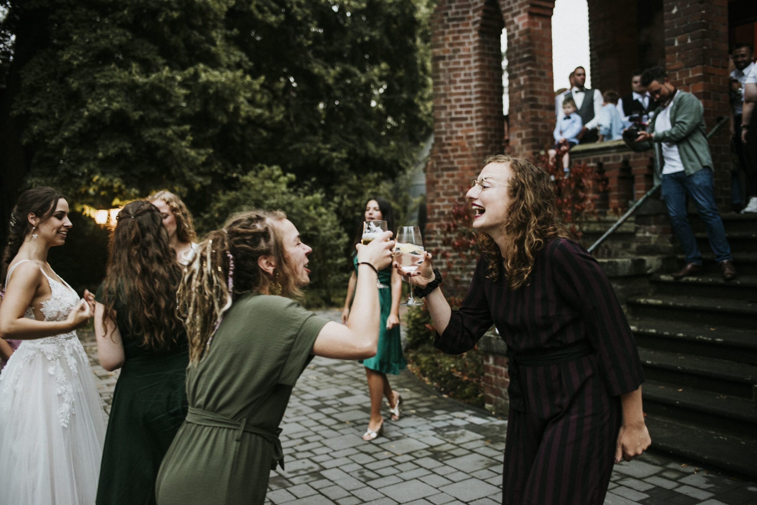 Group celebrating outdoors, person in wedding dress, two toasting with drinks, others on stairs.