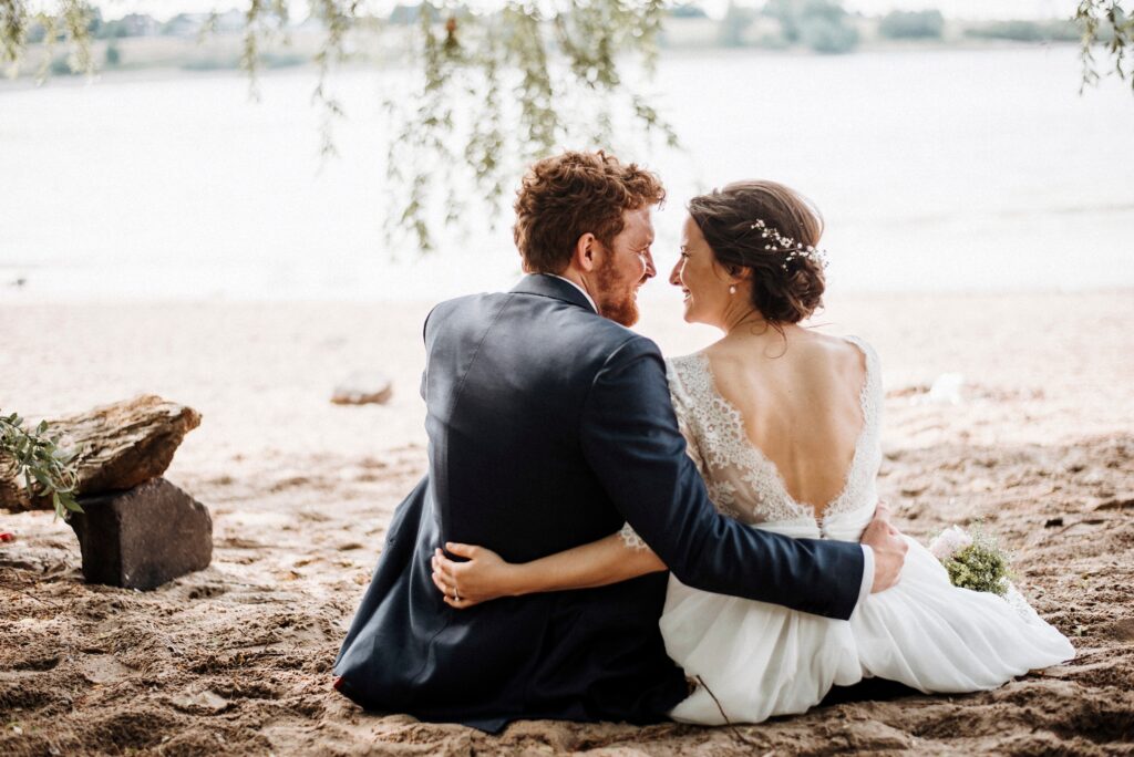 Paar in eleganter Kleidung sitzt eng am Strand, umgeben von Wasser, ruhige Atmosphäre.