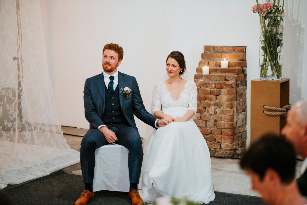 Paar in Hochzeitskleidung auf Stühlen, Kerzen und Blumen im Hintergrund, halten Hände.