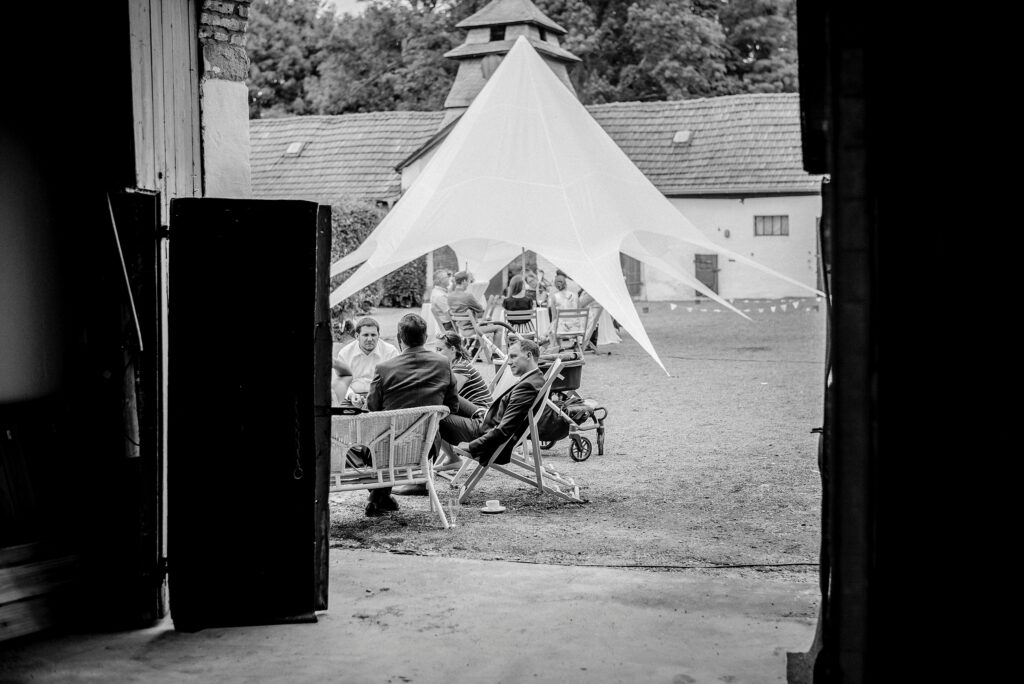 Menschen auf Stühlen unter weißem Zelt in einem Hof, Gebäude und Bäume im Hintergrund.