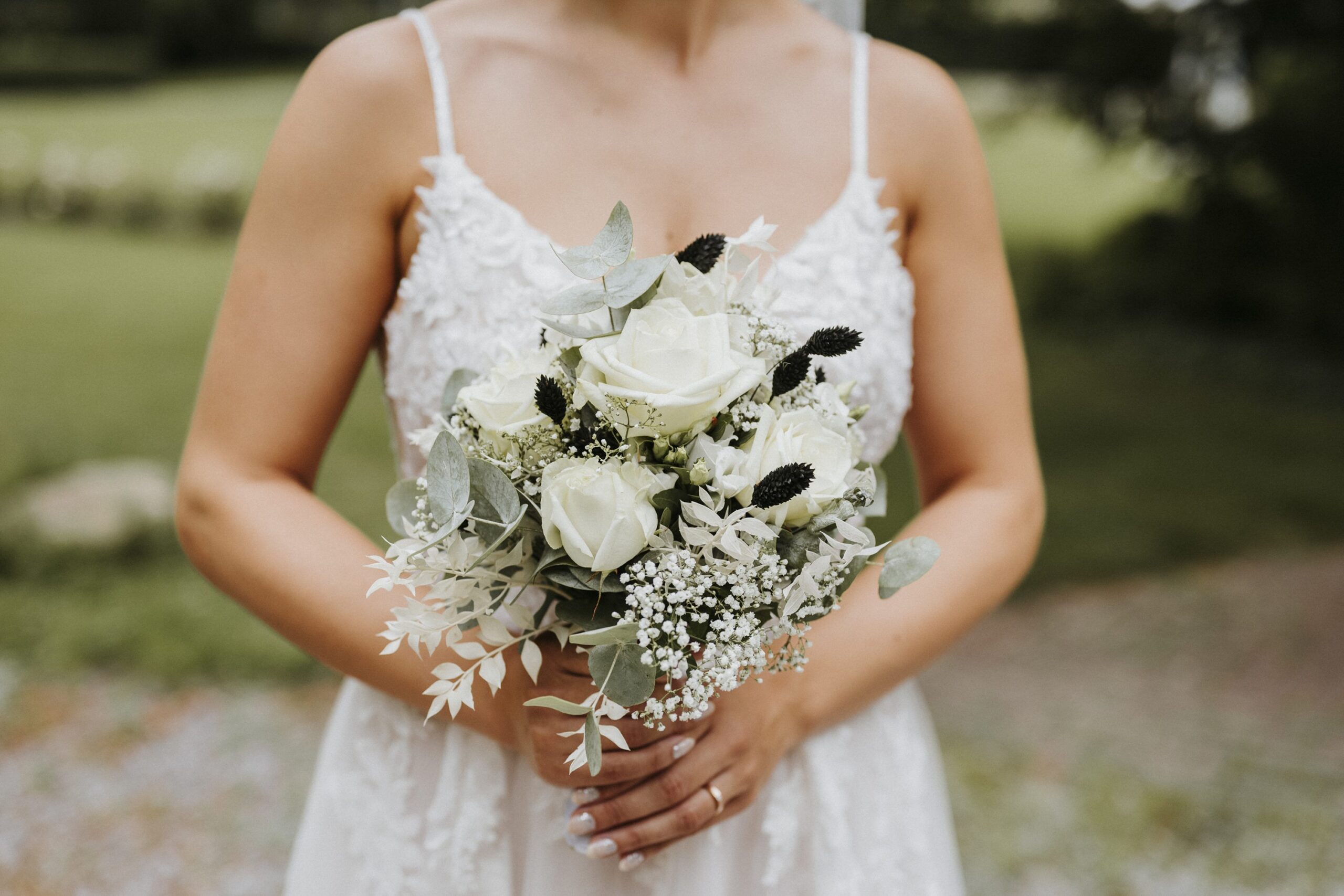Person in weißem Kleid hält Blumenstrauß mit weißen Rosen und Eukalyptus vor unscharfem Grün.