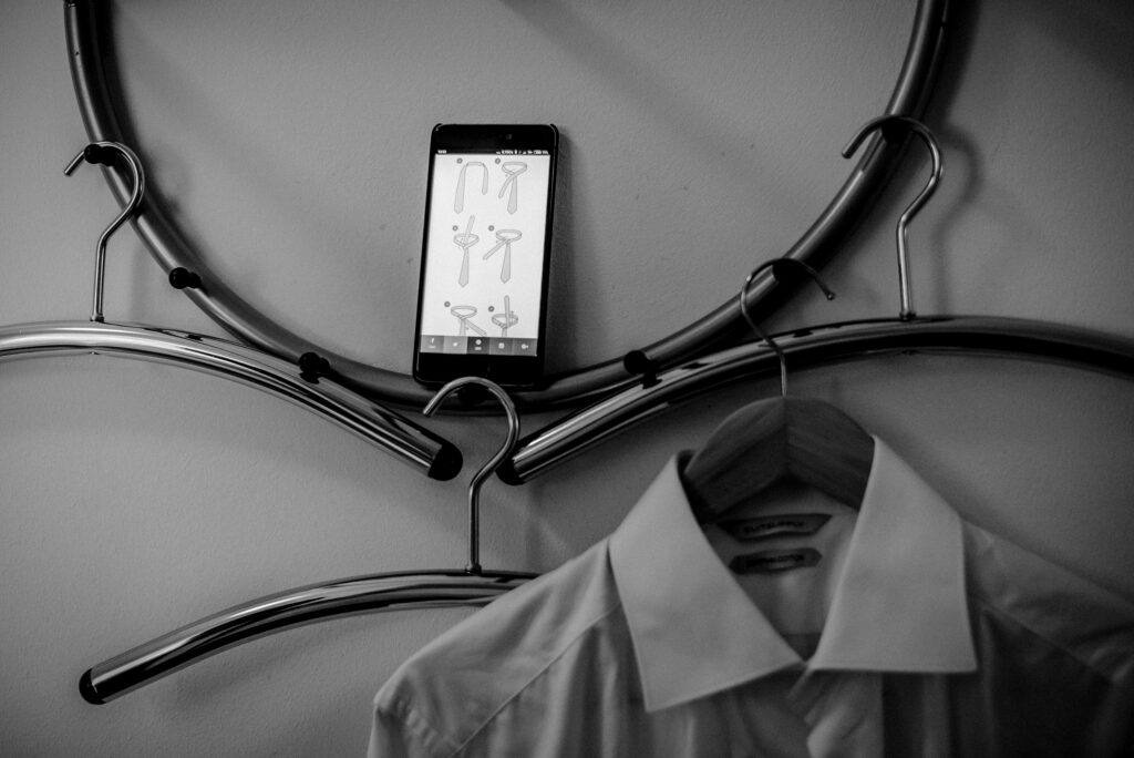 Smartphone mit Krawattenanleitungen an der Wand zwischen Kleiderbügeln, weißes Hemd darunter.