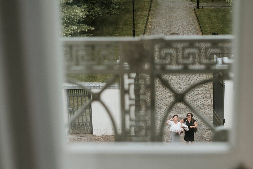 Blick durch Fenster auf zwei Personen, eine mit Blumenstrauß; dekoratives Gitter im Vordergrund.