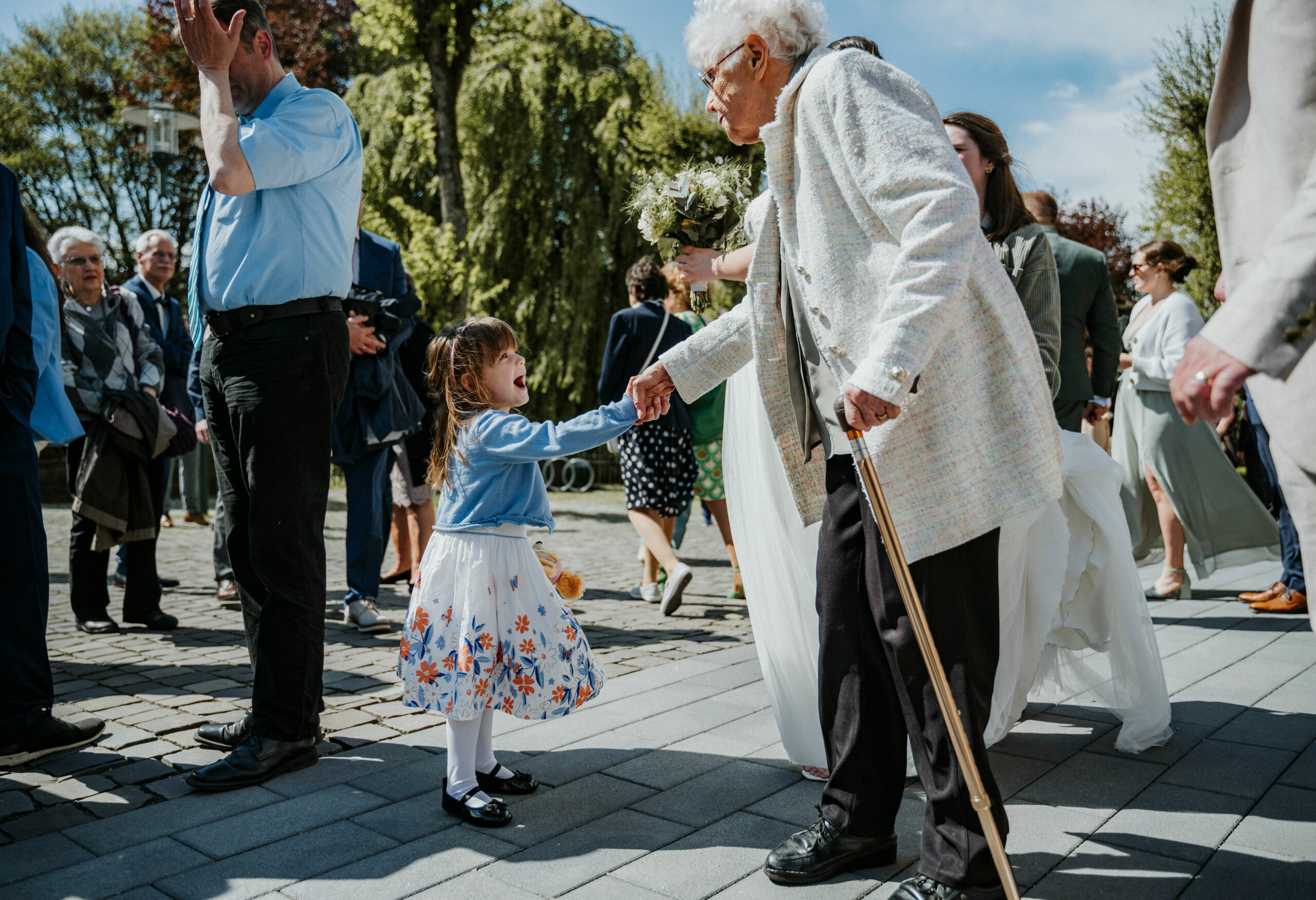 Oma und Mädchen halten Hände bei sonniger Versammlung im Freien auf gepflastertem Platz.