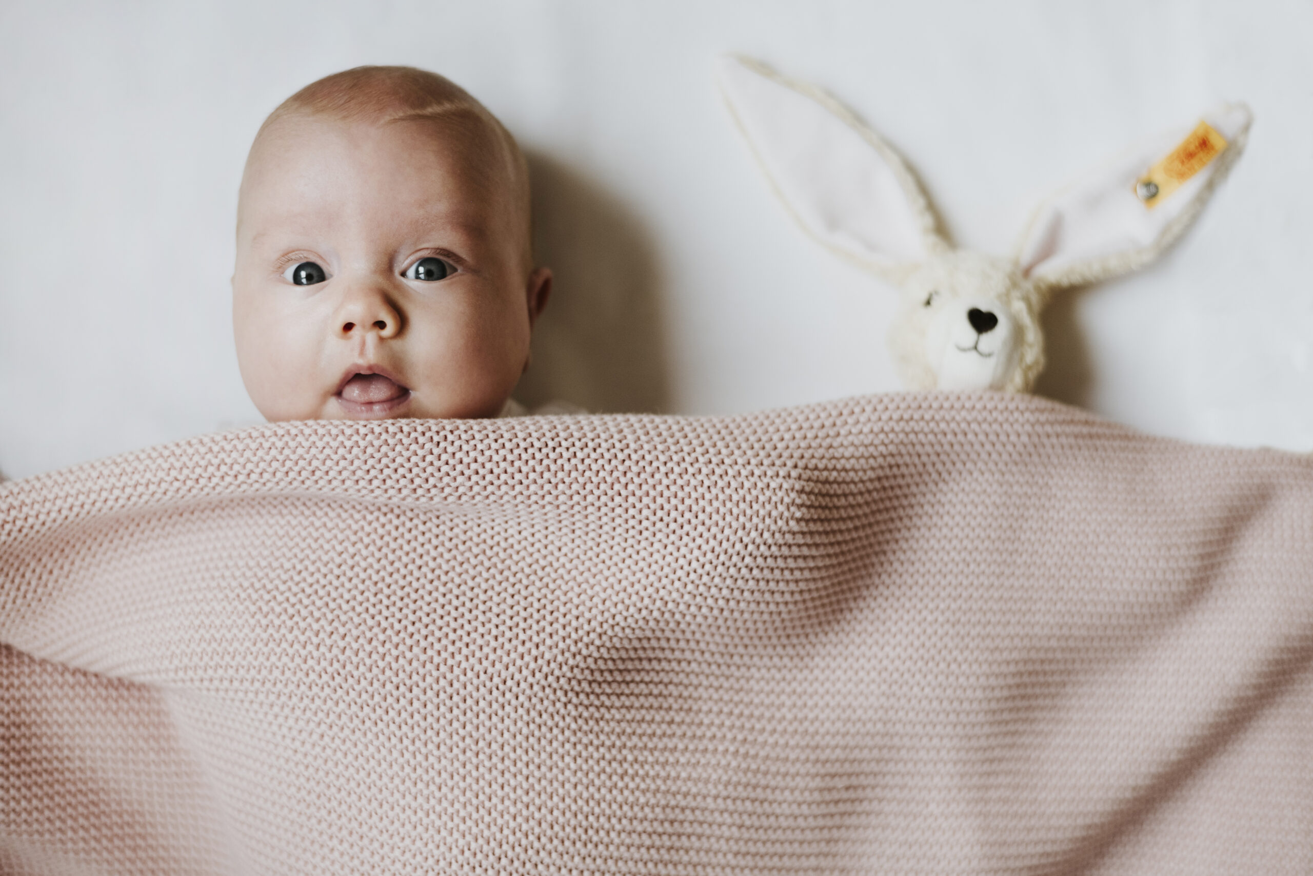 Baby liegt auf dem Rücken mit gestrickter Decke und Hasen-Plüschtier daneben.