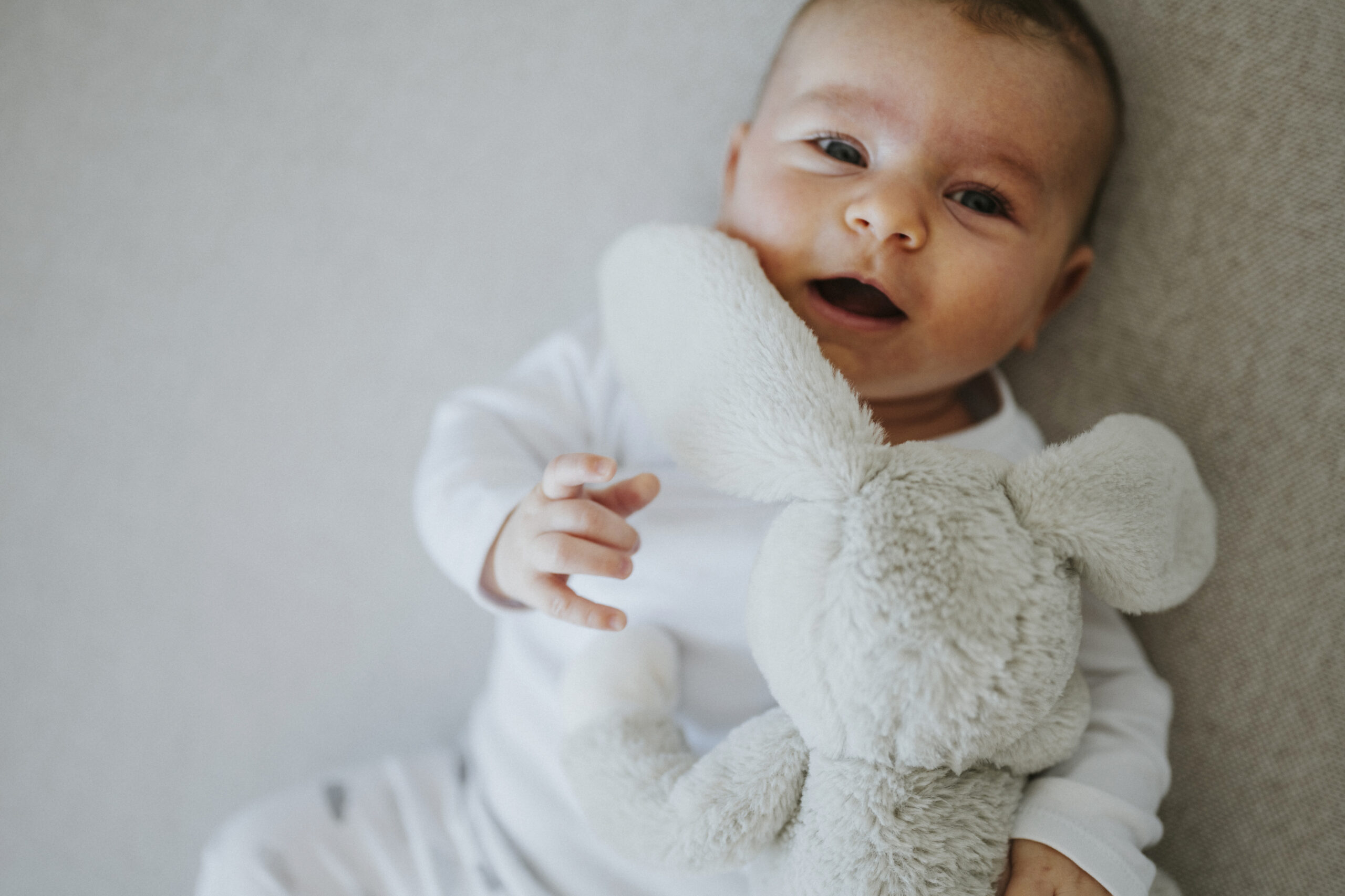 Ein Baby in weißem Strampler hält ein Plüschtier und liegt auf einem weichen Untergrund.