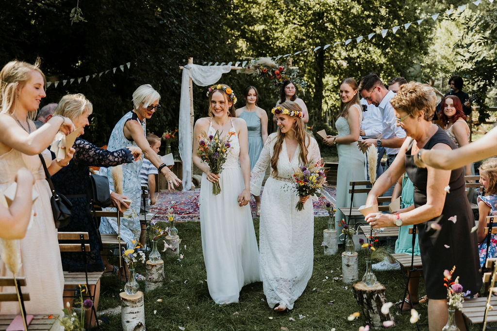 Zwei Bräute in weißen Kleidern bei einer grünen, festlichen Outdoor-Hochzeit mit Gästen.