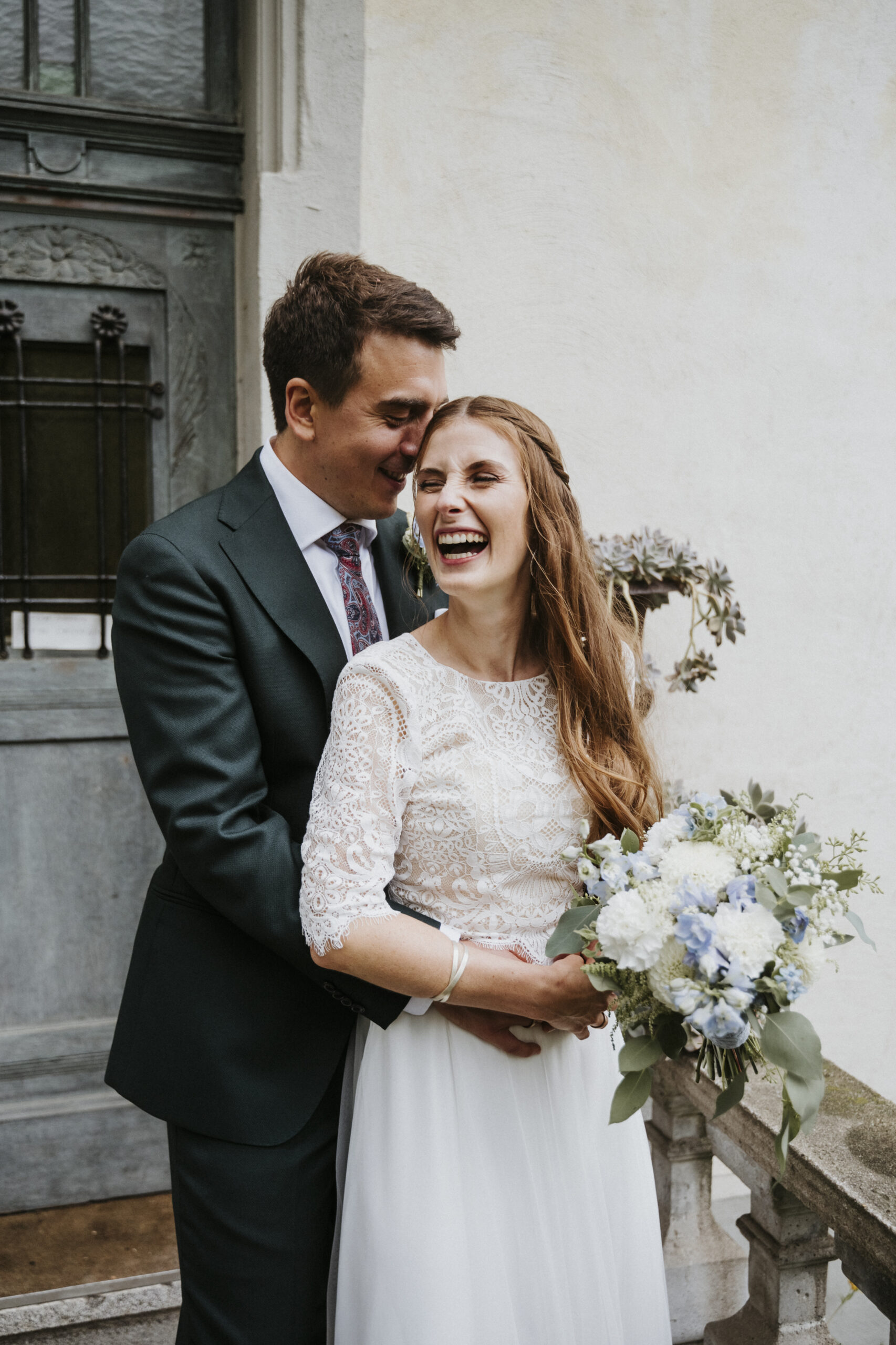 Paar posiert lächelnd vor Tür; Frau im weißen Kleid mit Blumenstrauß, Mann in dunklem Anzug.
