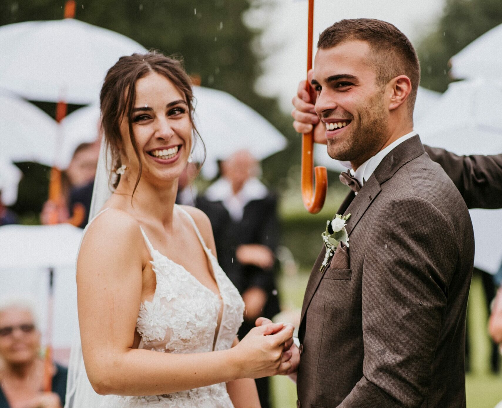 Brautpaar lächelt unter weißem Regenschirm, Brautkleid mit Spitze, Bräutigam im Anzug.