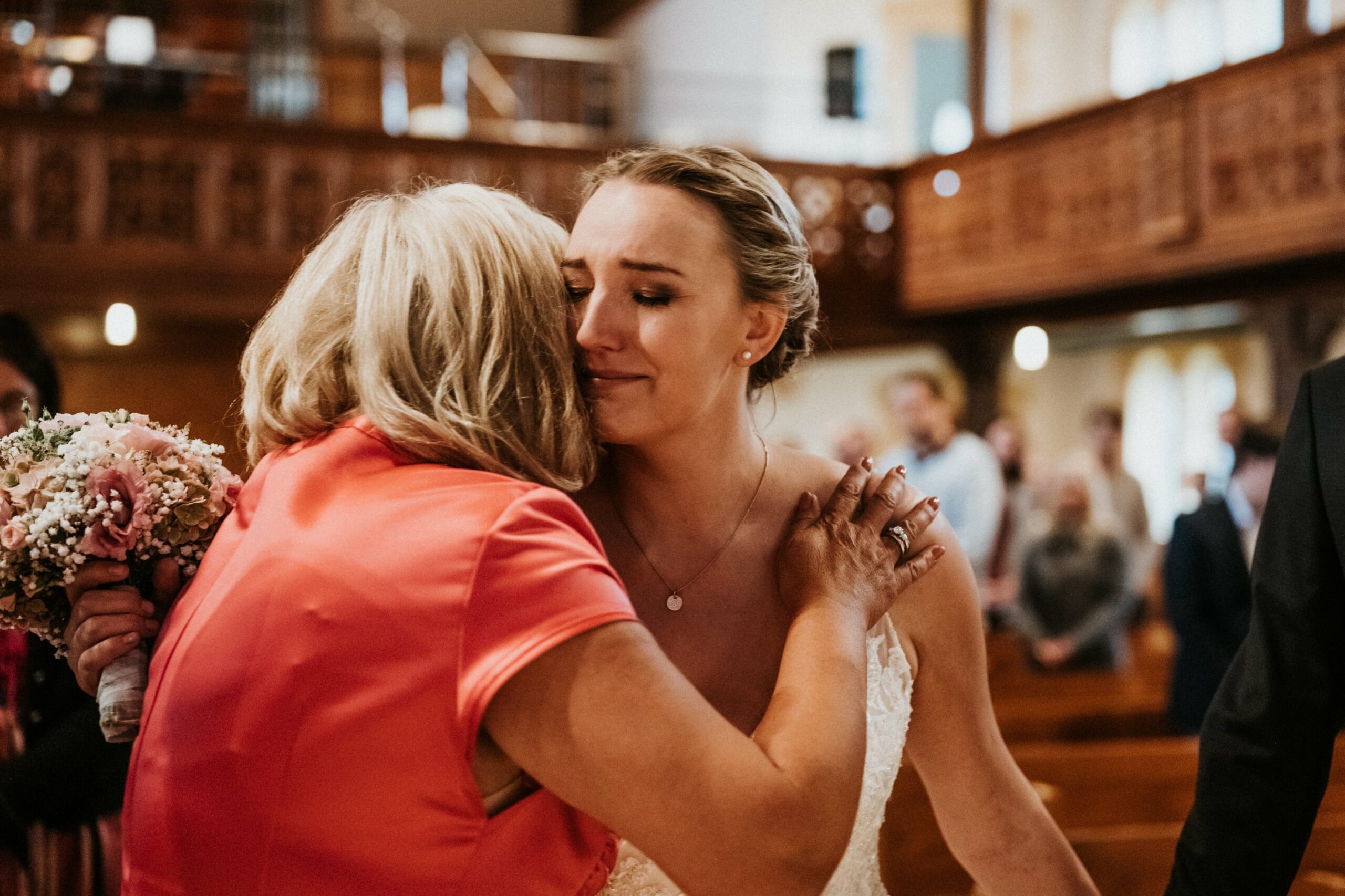 Die Braut umarmt ihre Mutter sehr emotional mit einem Blumenstrauß in einer Kirchenumgebung.