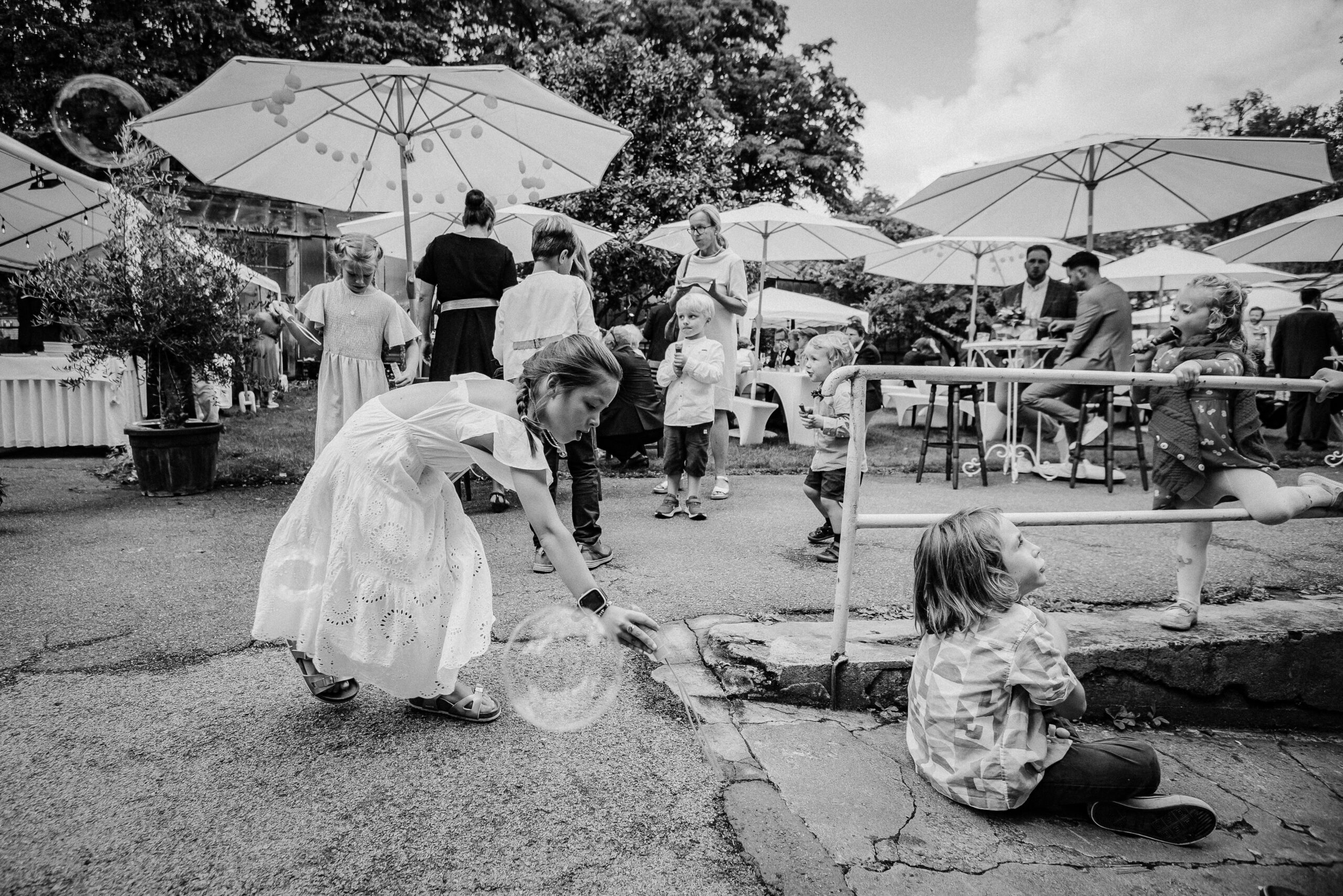 Kinder spielen draußen unter Sonnenschirmen, fangen Seifenblasen, Erwachsene im Hintergrund.