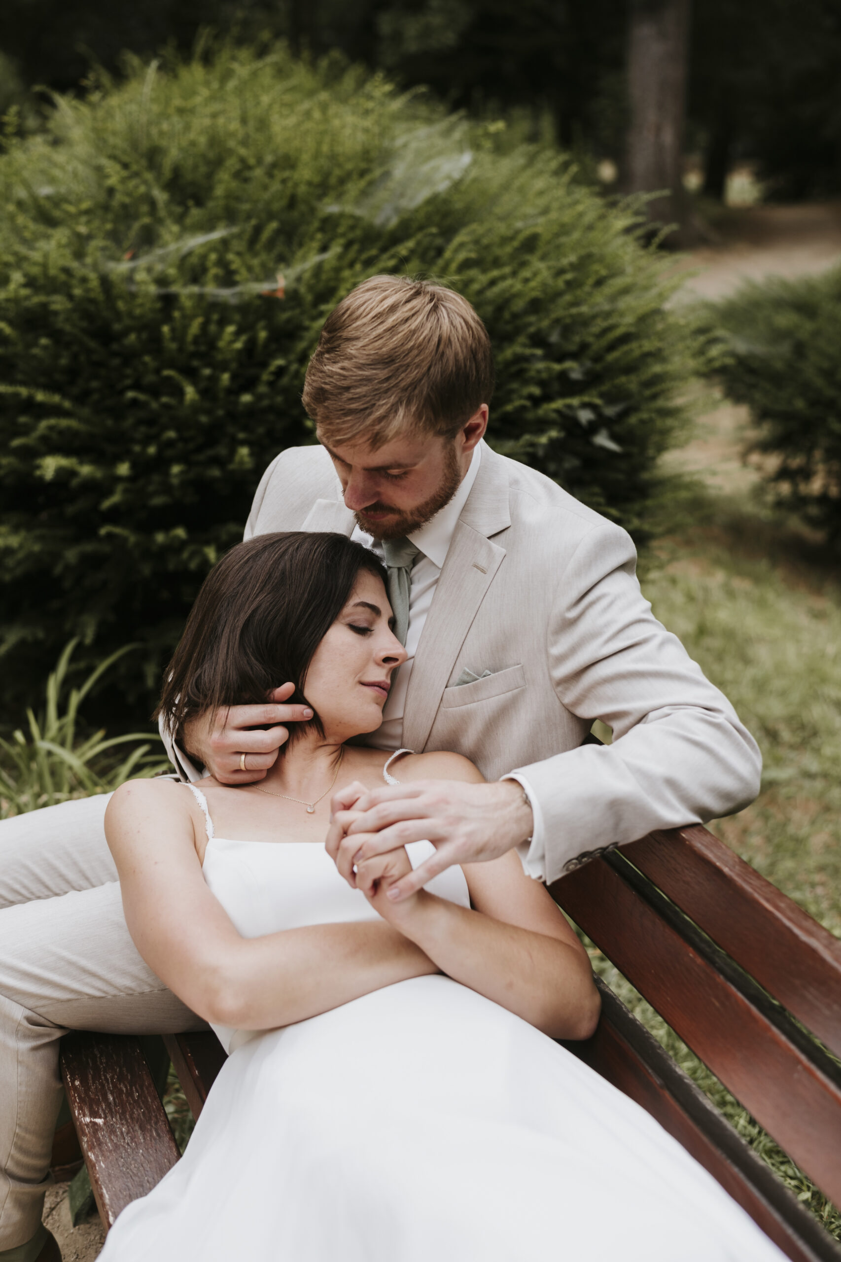 Couple in park on bench, man embraces woman in white dress, surrounded by greenery.