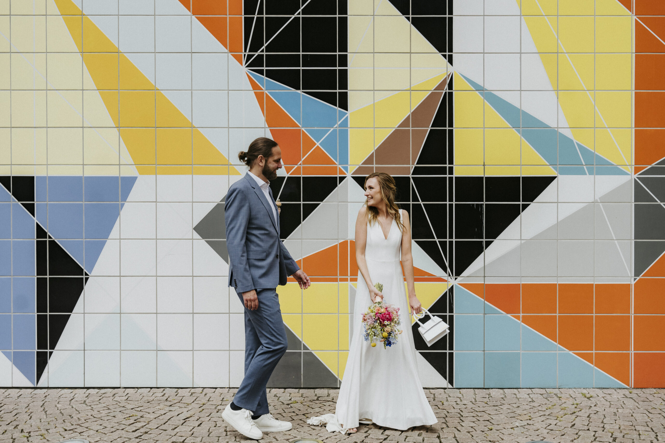 Paar in eleganter Kleidung vor bunter geometrischer Wand auf Kopfsteinpflaster stehend.