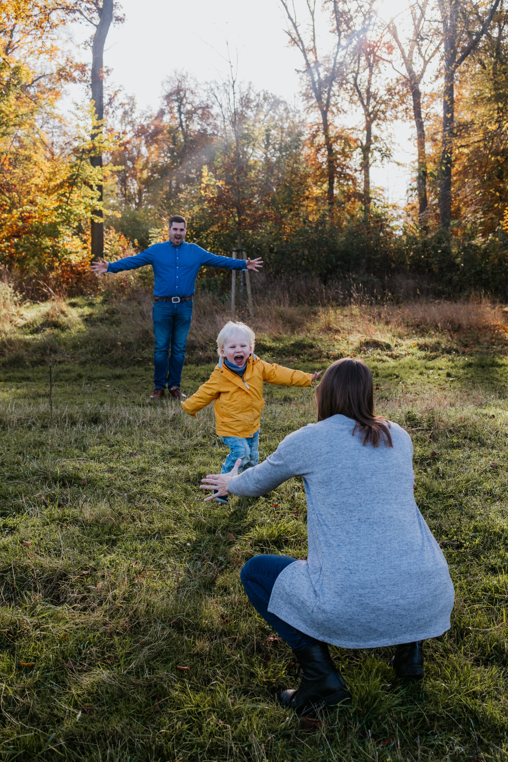 Familie in herbstlichem Wald, Kind läuft zu Erwachsenen, Laub und buntes Laub im Hintergrund.