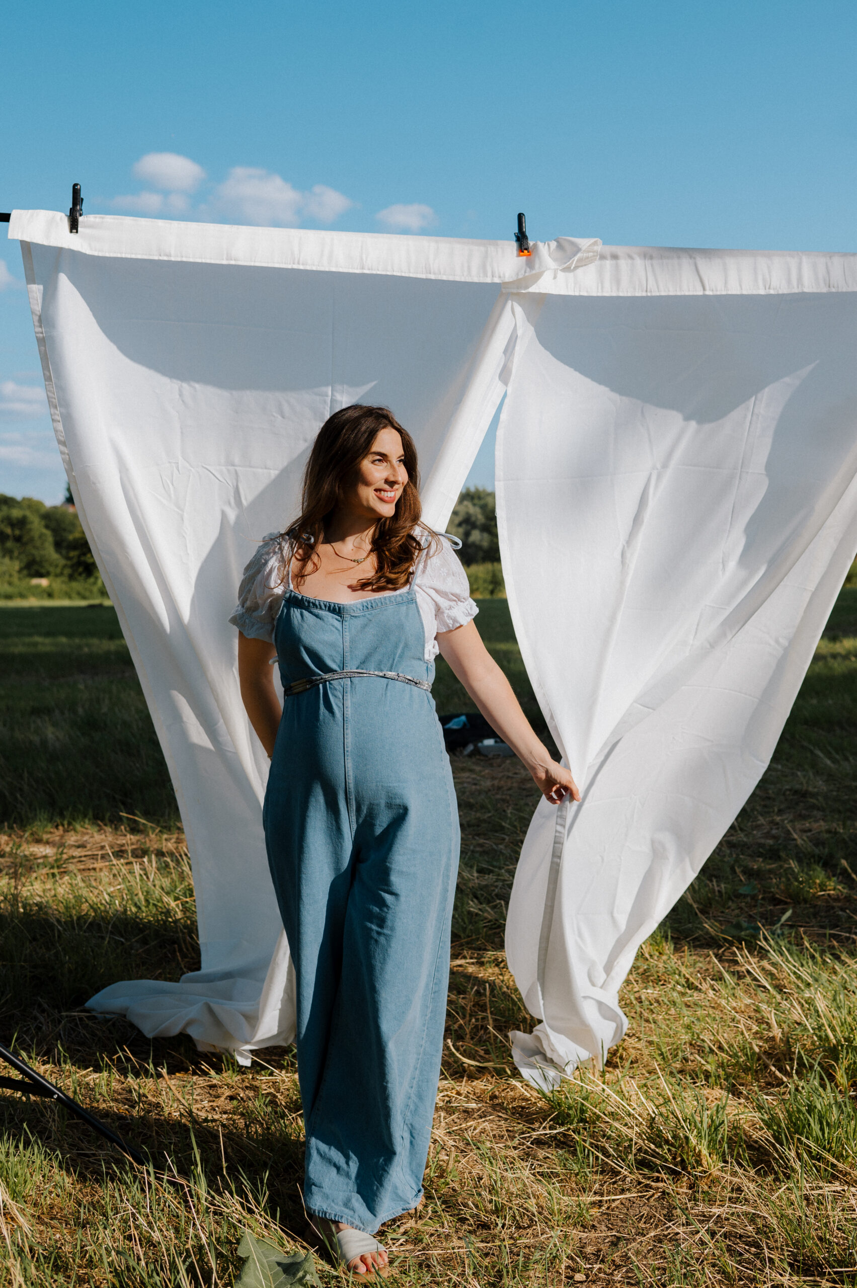 Frau in blauem Jumpsuit vor weißem Vorhang, grüne Wiese und sonniges Wetter im Hintergrund.