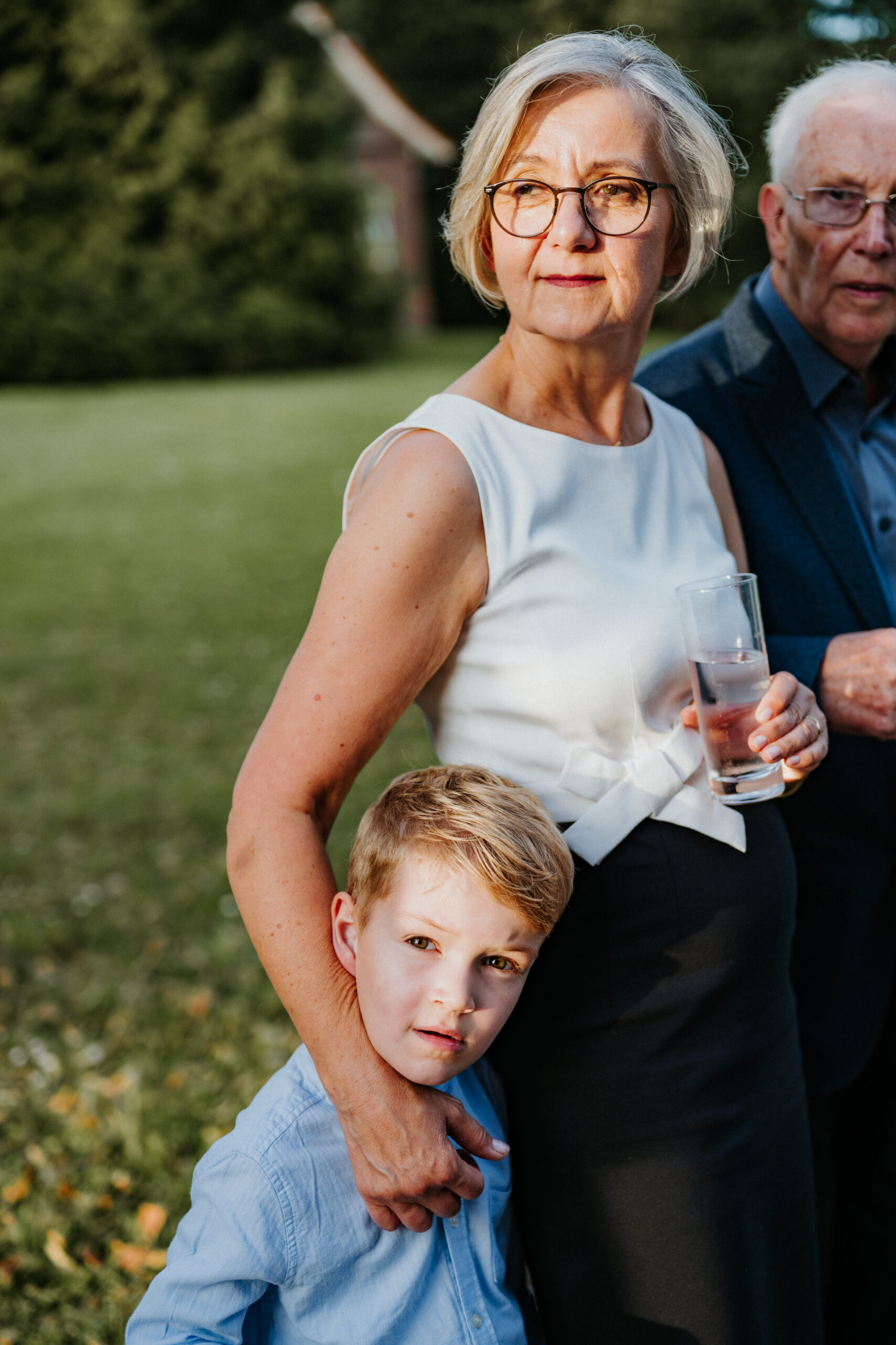 Frau mit Brille hält ein Glas, steht neben Junge im blauen Hemd, grüne Landschaft im Hintergrund.