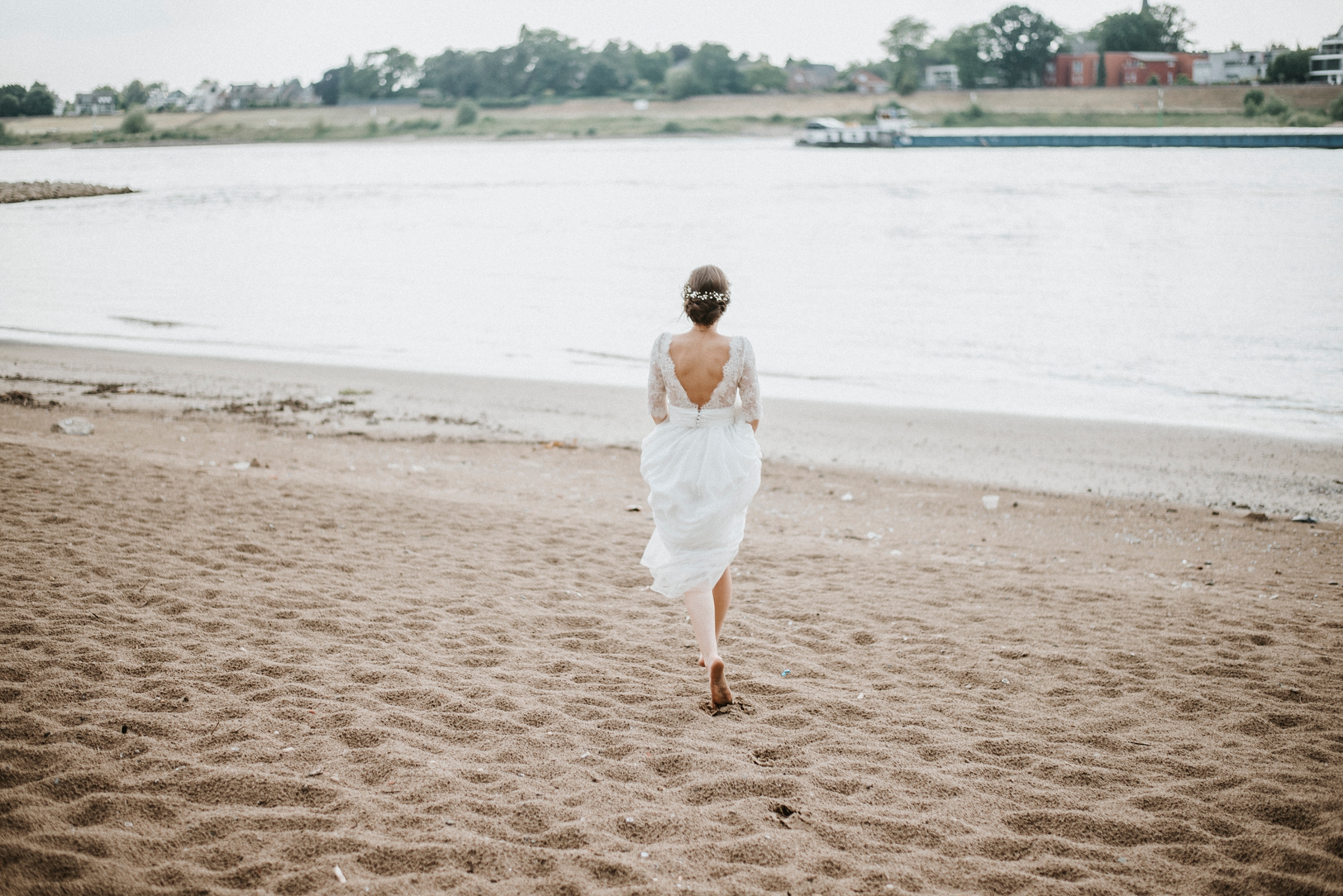 Frau im weißen Kleid barfuß am Sandstrand, blickt aufs Wasser, Flusslandschaft im Hintergrund.