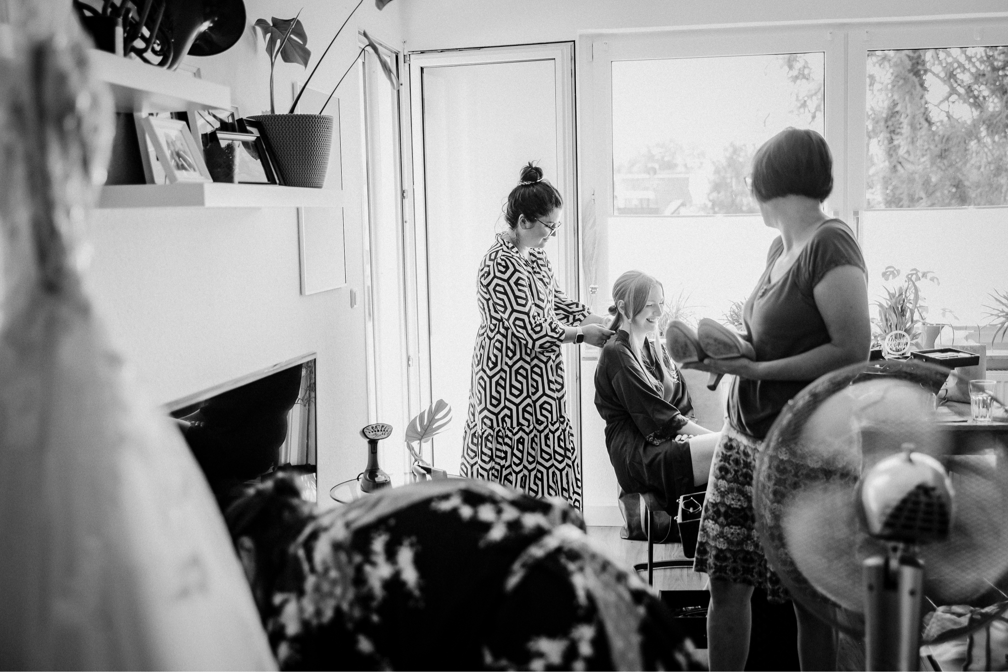 Zwei Frauen frisieren eine sitzende Frau, eine hält Schuhe, im Raum mit Pflanzen und Fenstern.